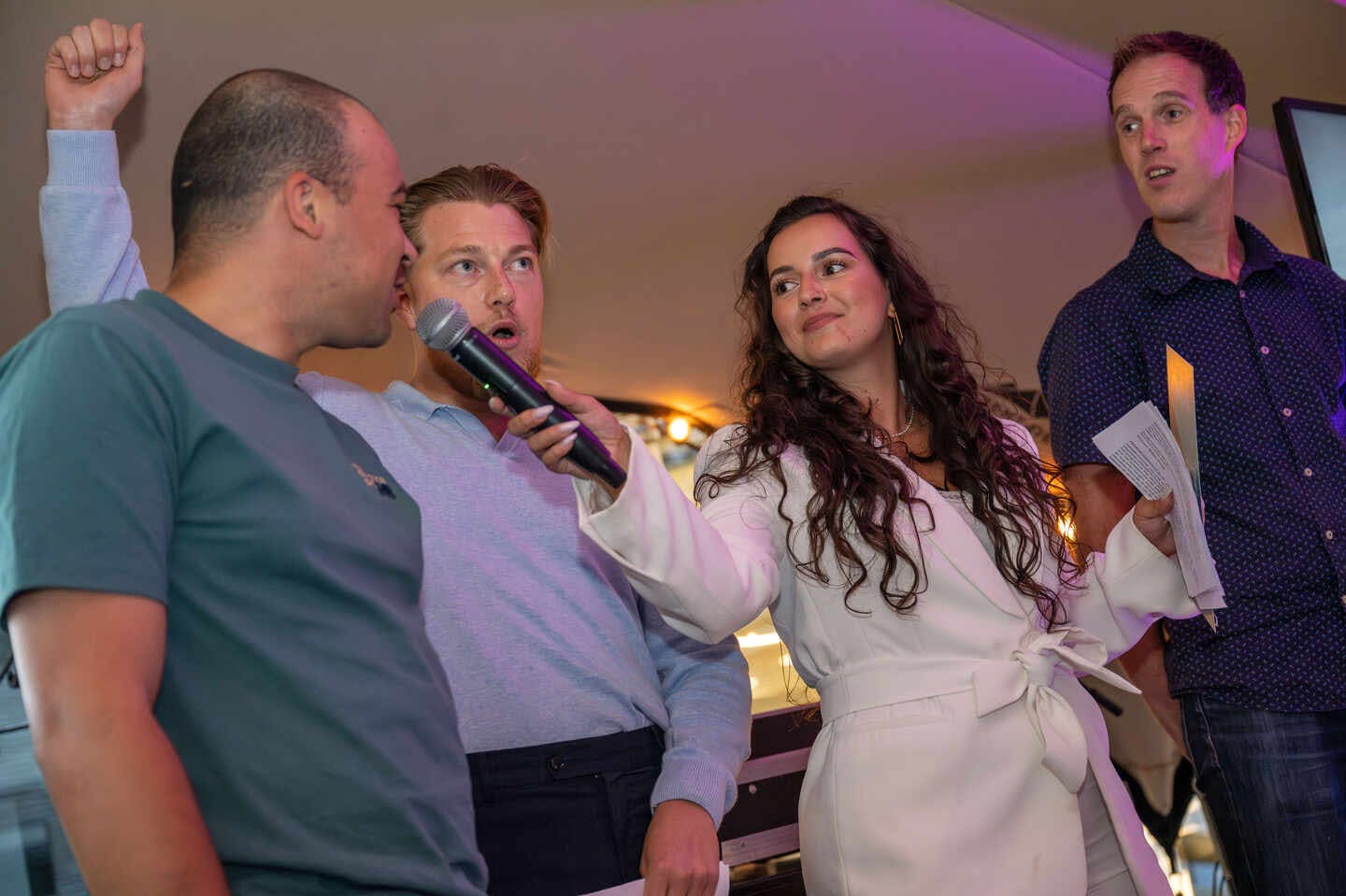 Tara op het podium tijdens het The.NextGen zomer feest met microfoon in hand. Drie NextGenners staan naast haar, waarvan er eentje in de microfoon praat .