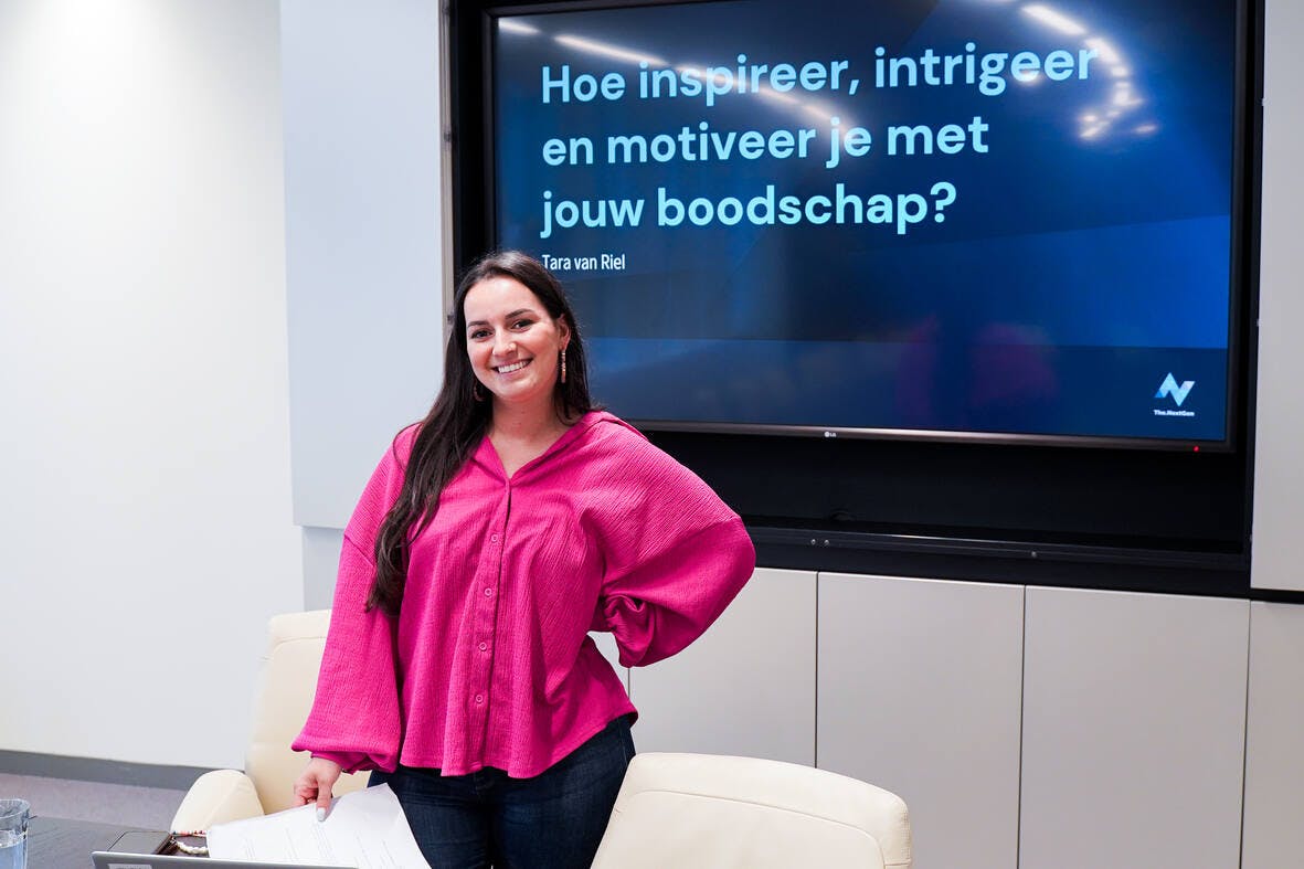 Tara staat achter een tafel, blaadje in haar hand, en lacht in de camera.