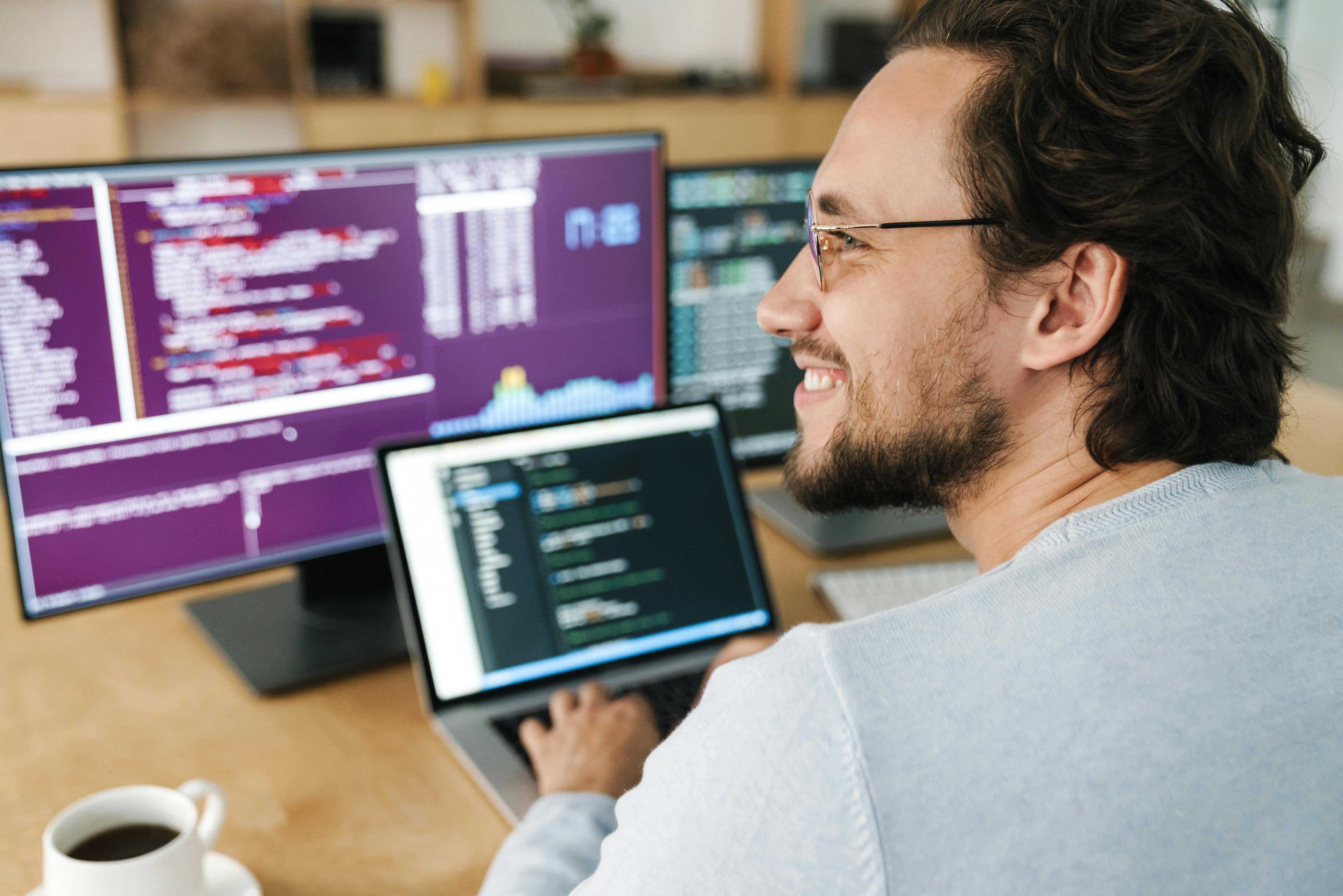 Smiling man with glasses in front of a laptop and 2 screens. Code displayed on all screens.