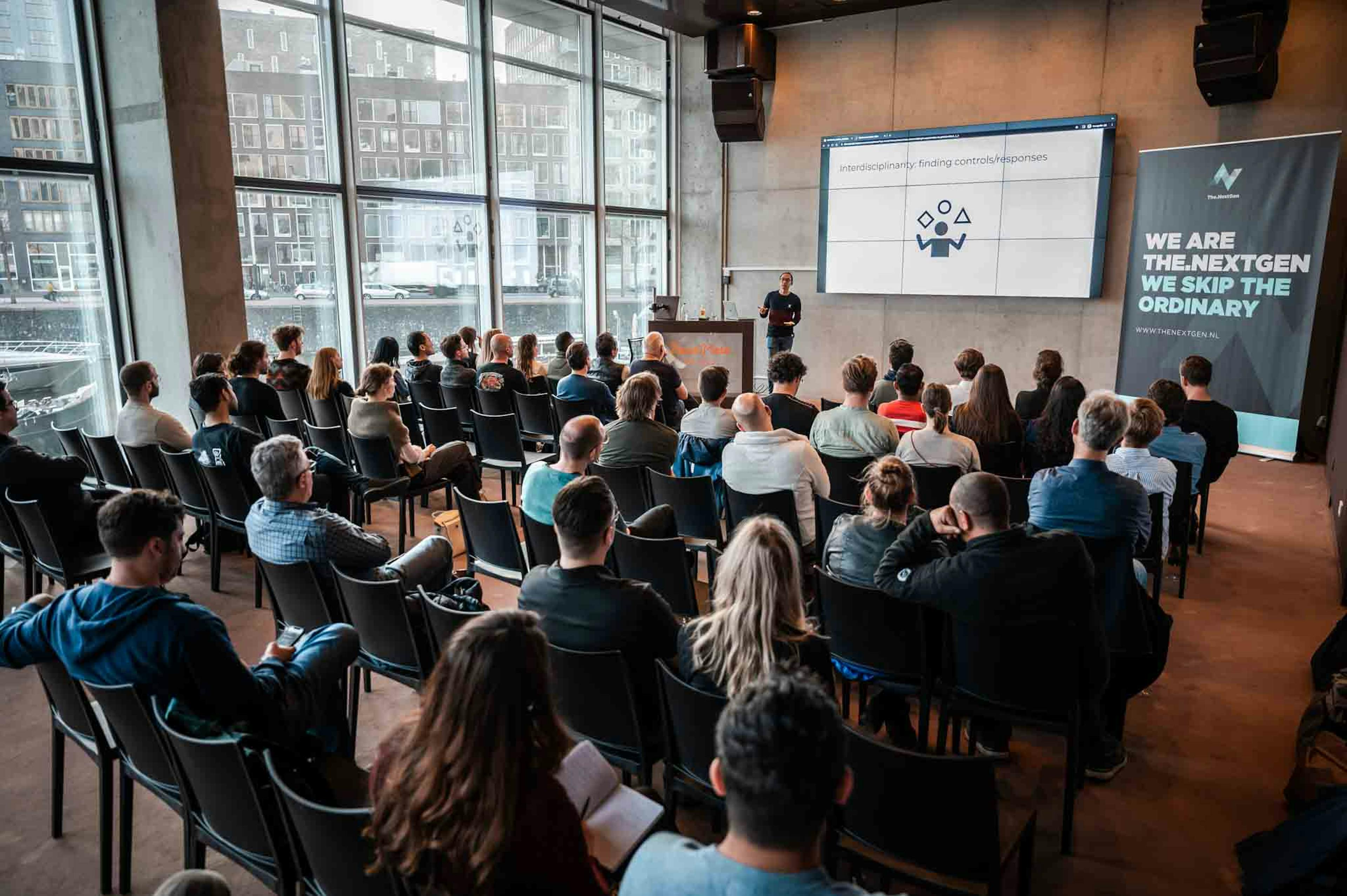 Image of an event room filled with people. There is one speaker in the front of the room presenting to the crowd, who are sitting in chairs. A The.NextGen banner is displayed in the top right.