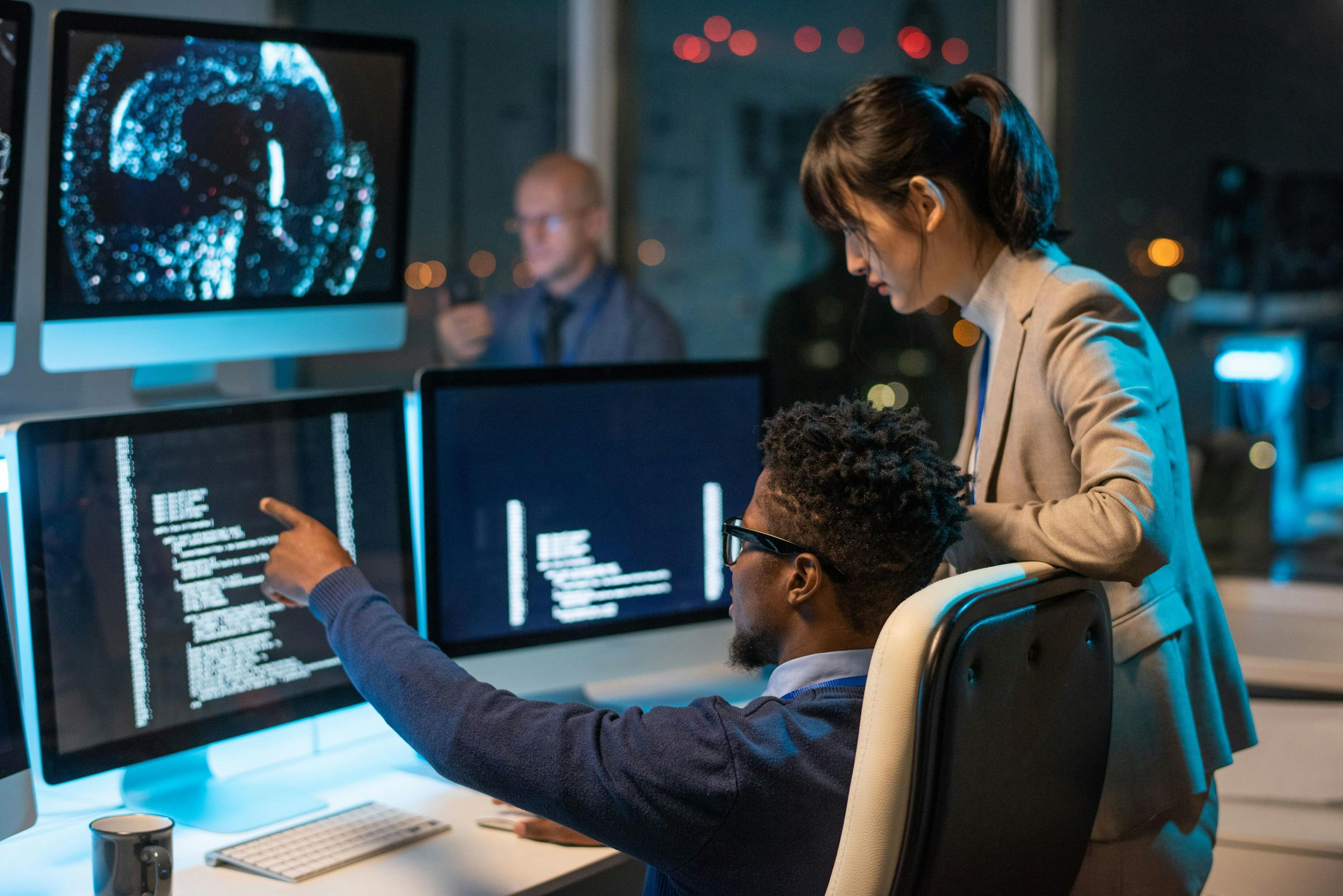 Two developers are looking at a screen. One is seated in a chair and is pointing towards the screen. On the screen code is displayed that both are looking at. 