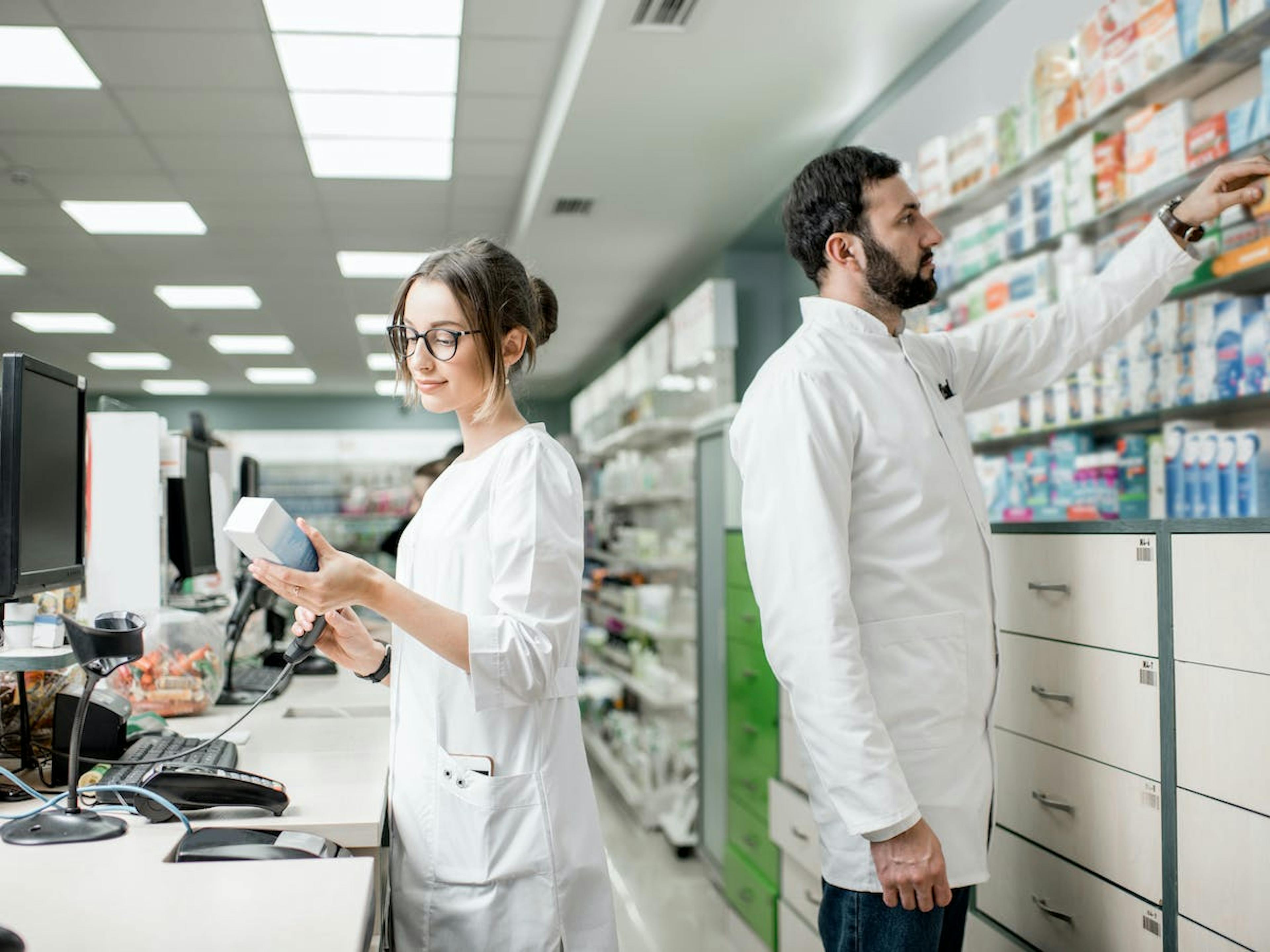 Twee apothekers staan in hun apotheek. De ene scant een item terwijl de ander iets uit een rij pakt. 