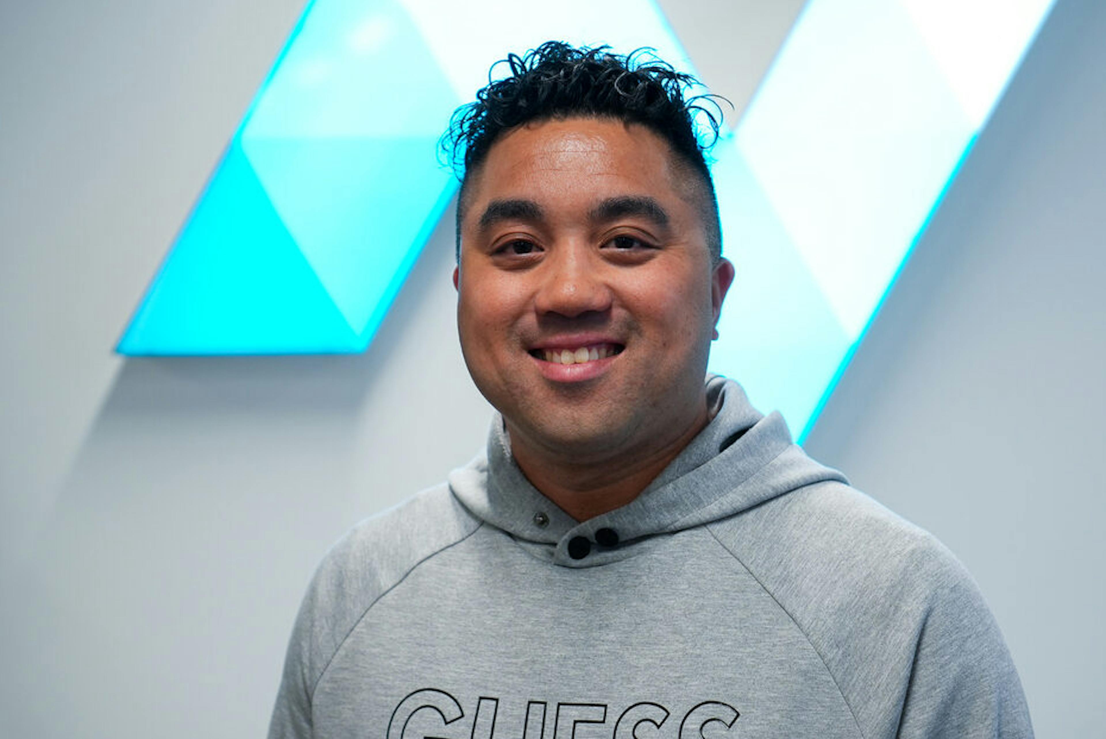 Richard Cepeda kijkt lachend in de camera terwijl hij voor het logo van The.NextGen staat. 