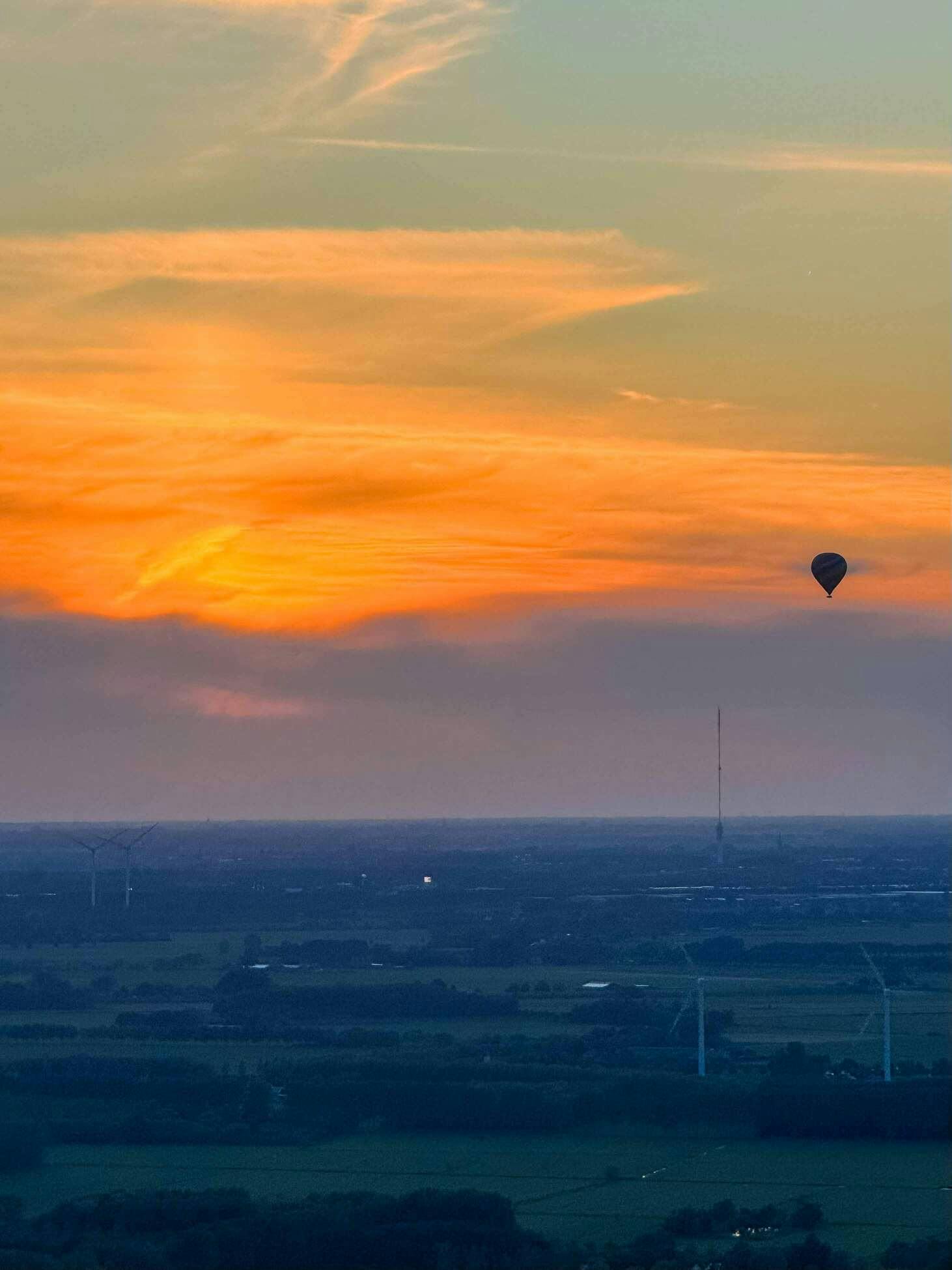 Een luchtballon is hoog in de lucht te zien als silhouette tegen de ondergaande zon boven het Nederlandse landschap.