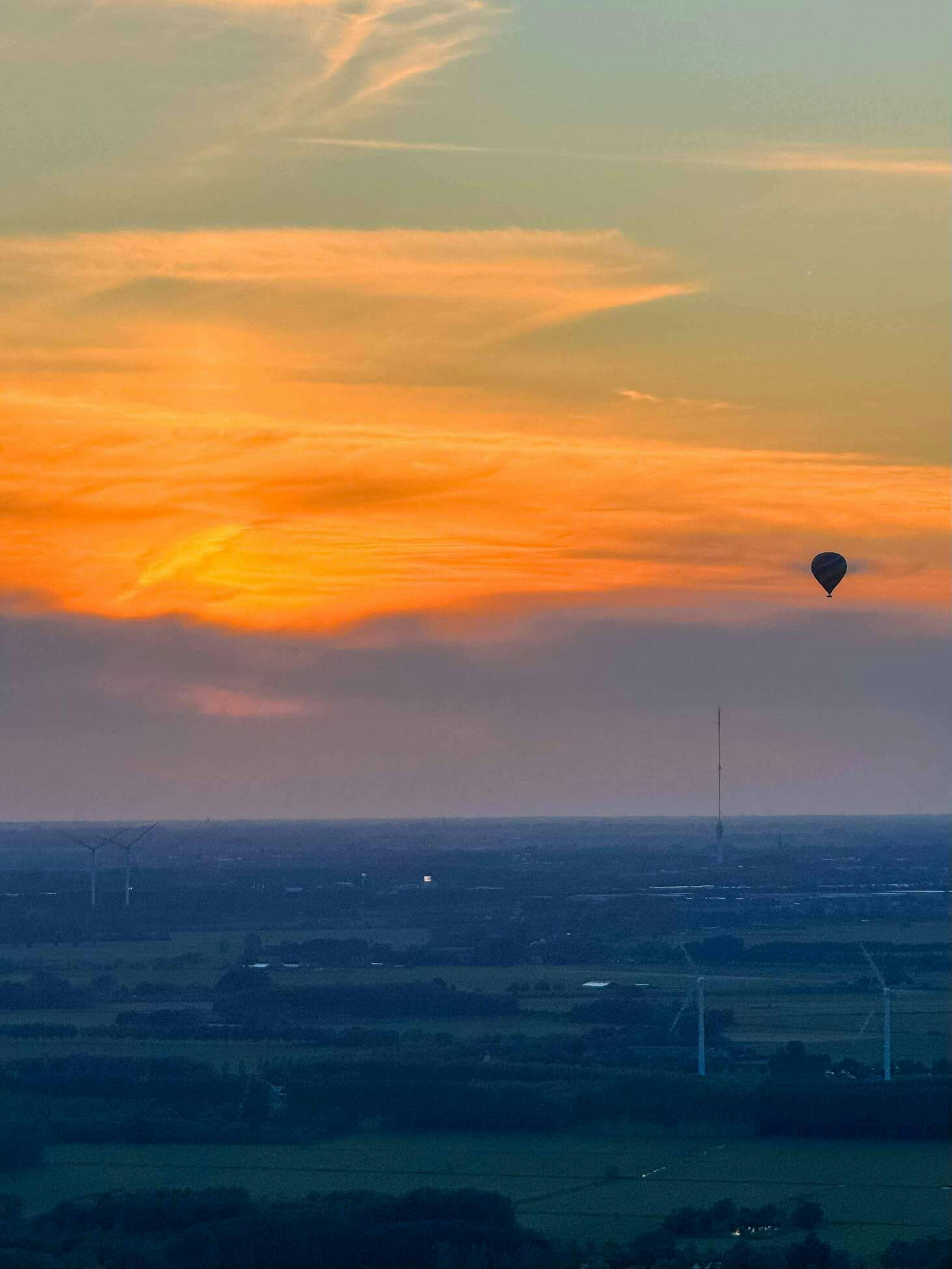 Een luchtballon is hoog in de lucht te zien als silhouette tegen de ondergaande zon boven het Nederlandse landschap. 
