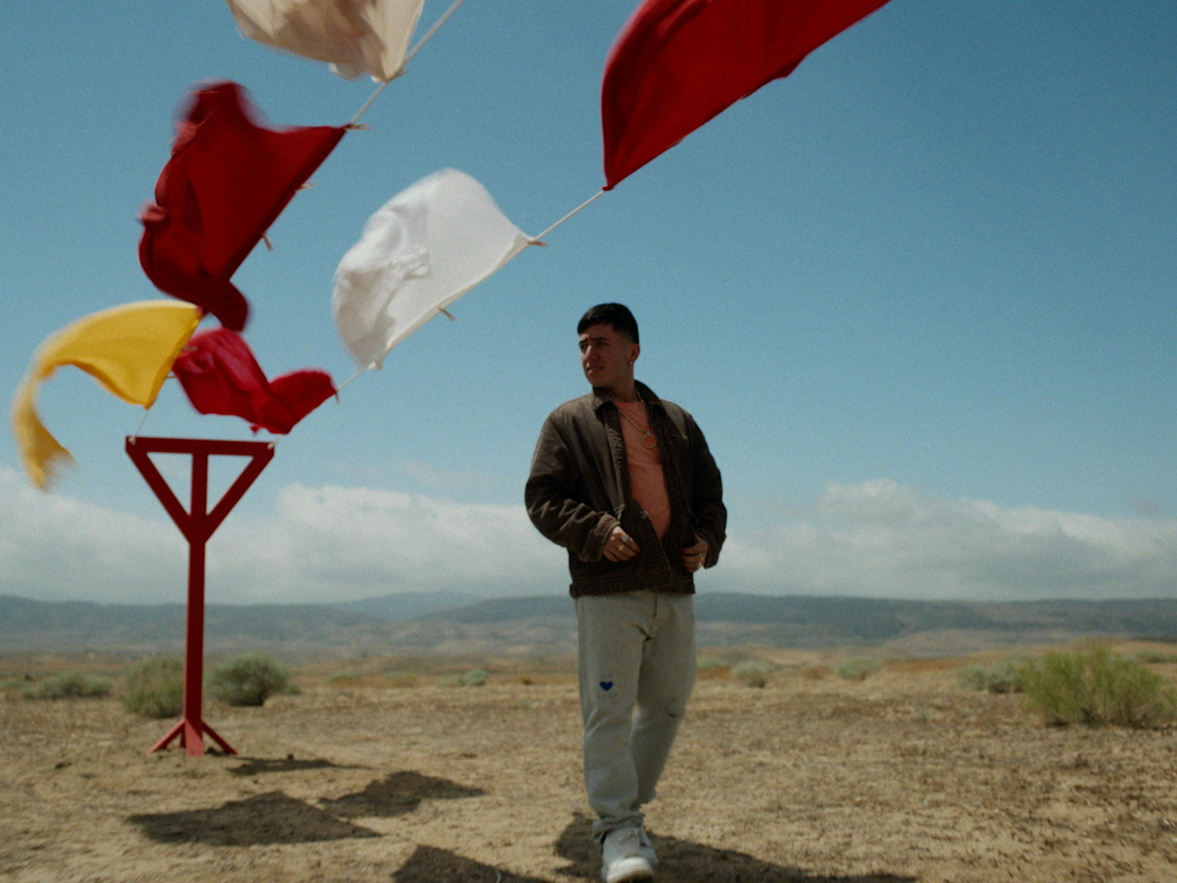 A man stands under a clotheslines in the desert with colorful cloths flowing in the wind