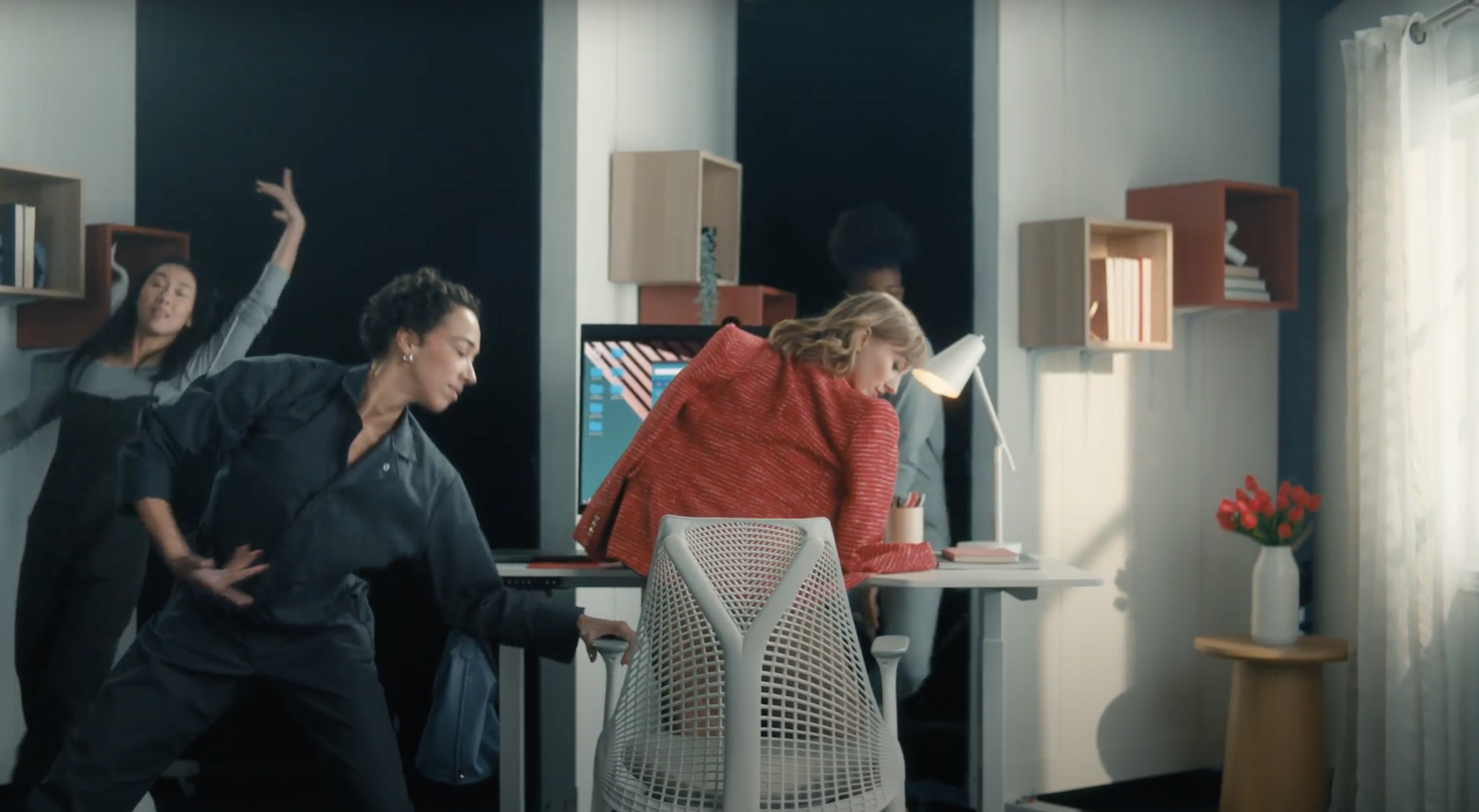 A woman stands in front of her desk, getting ready to dance while two other dancers appear beside her