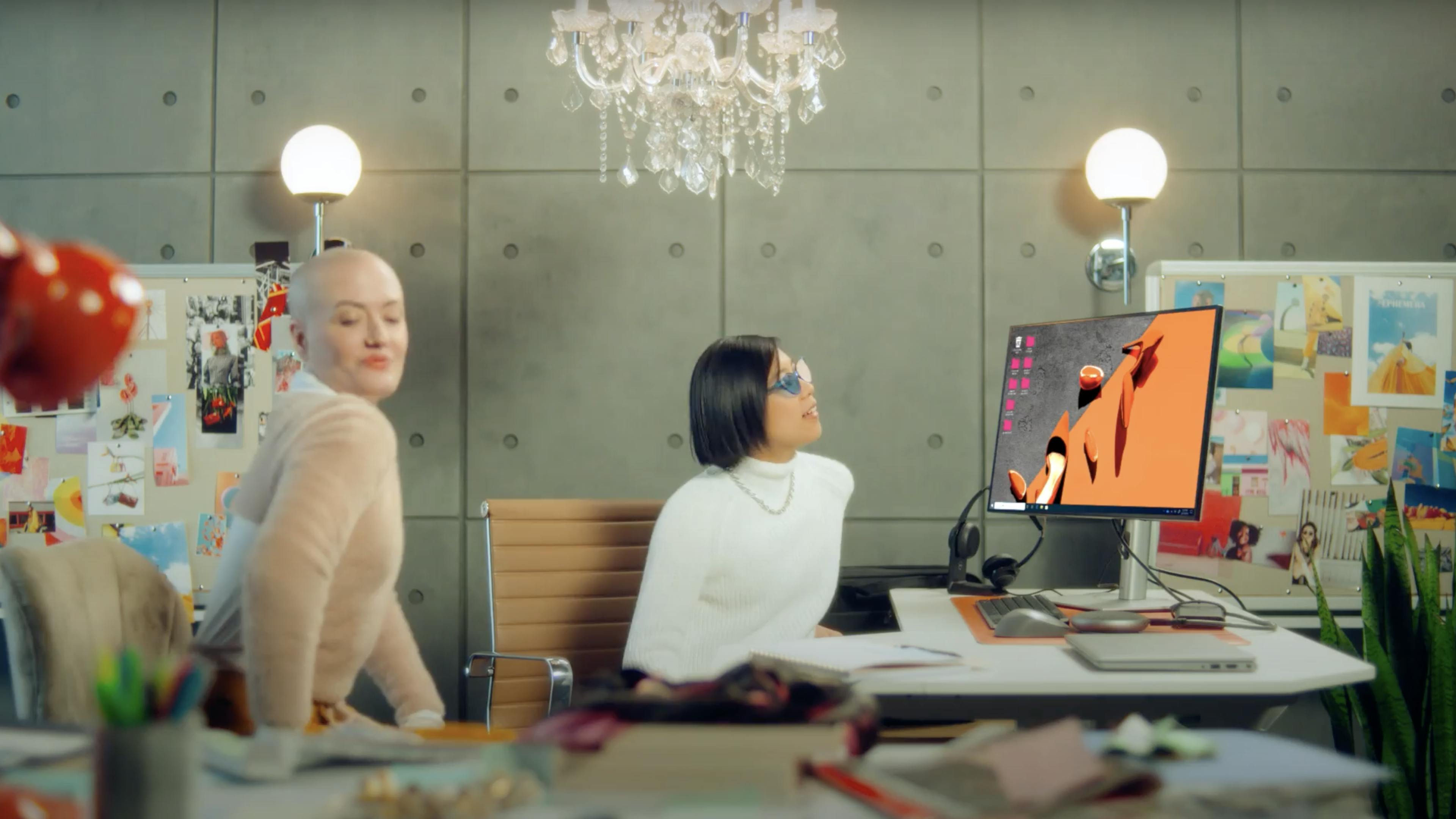 A woman with sunglasses looks at her computer monitor while another woman dances beside her