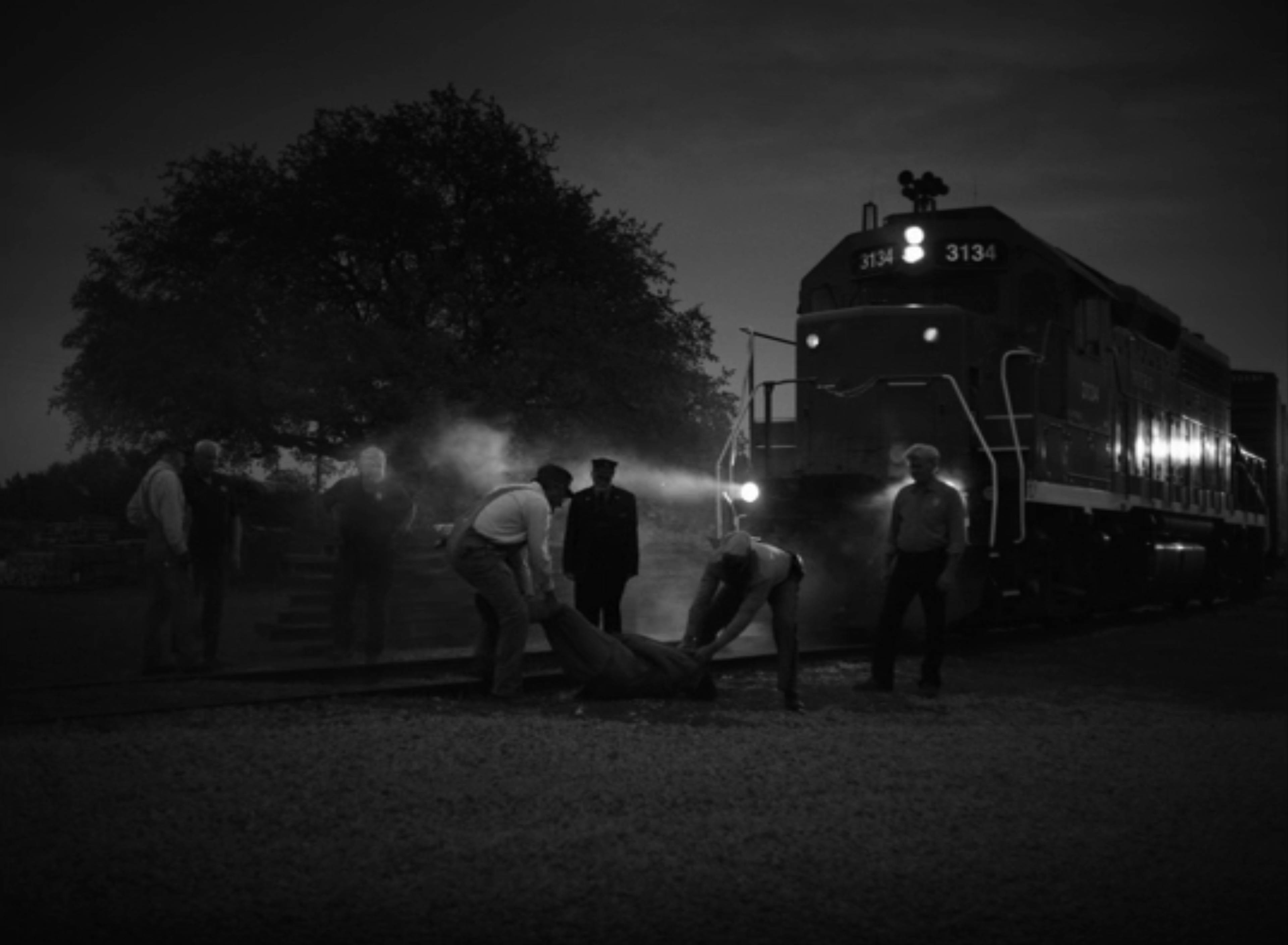 A black and white image of train workers dragging something off the tracks and a train right behind them