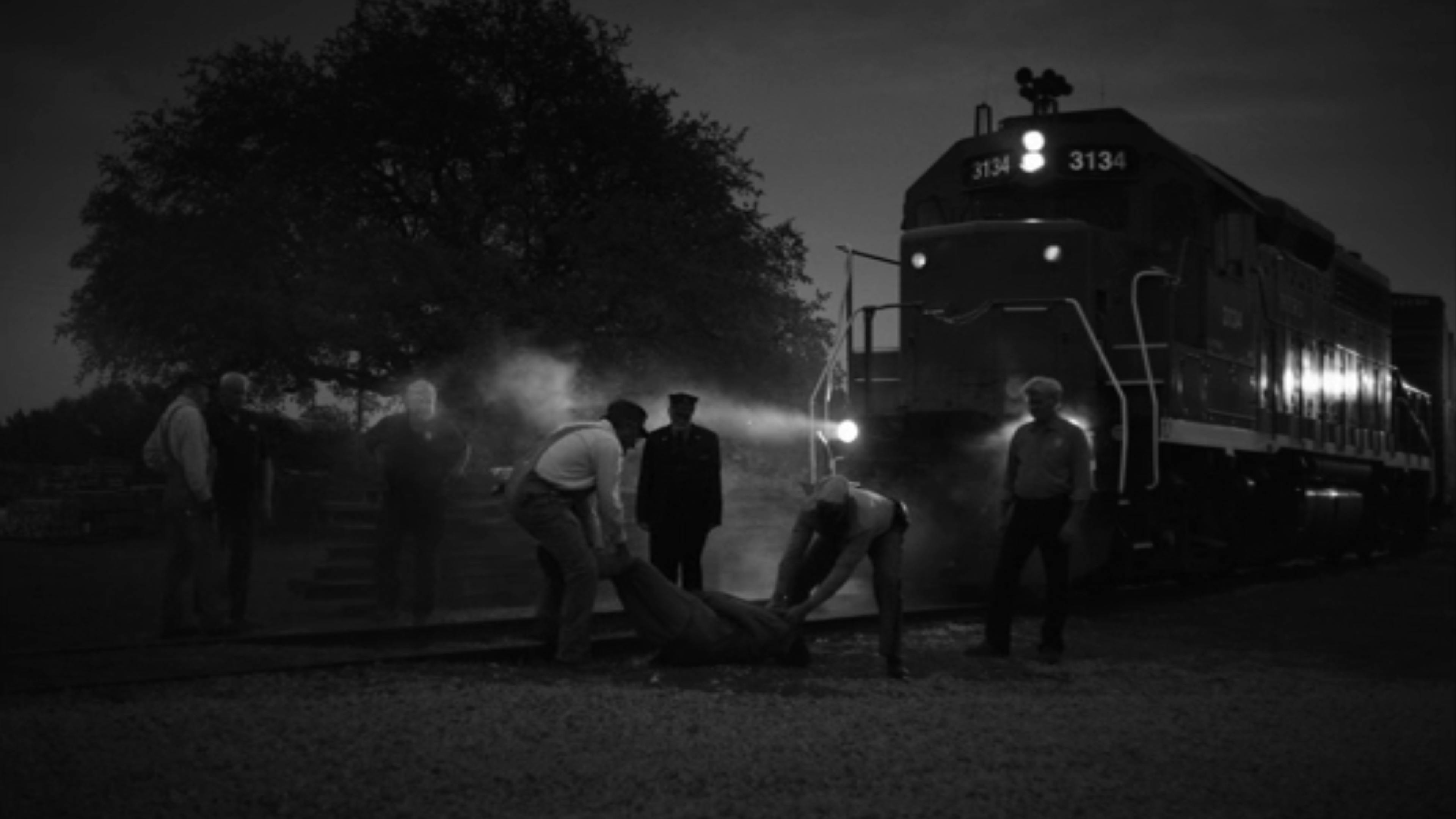 A black and white image of train workers dragging something off the tracks and a train right behind them