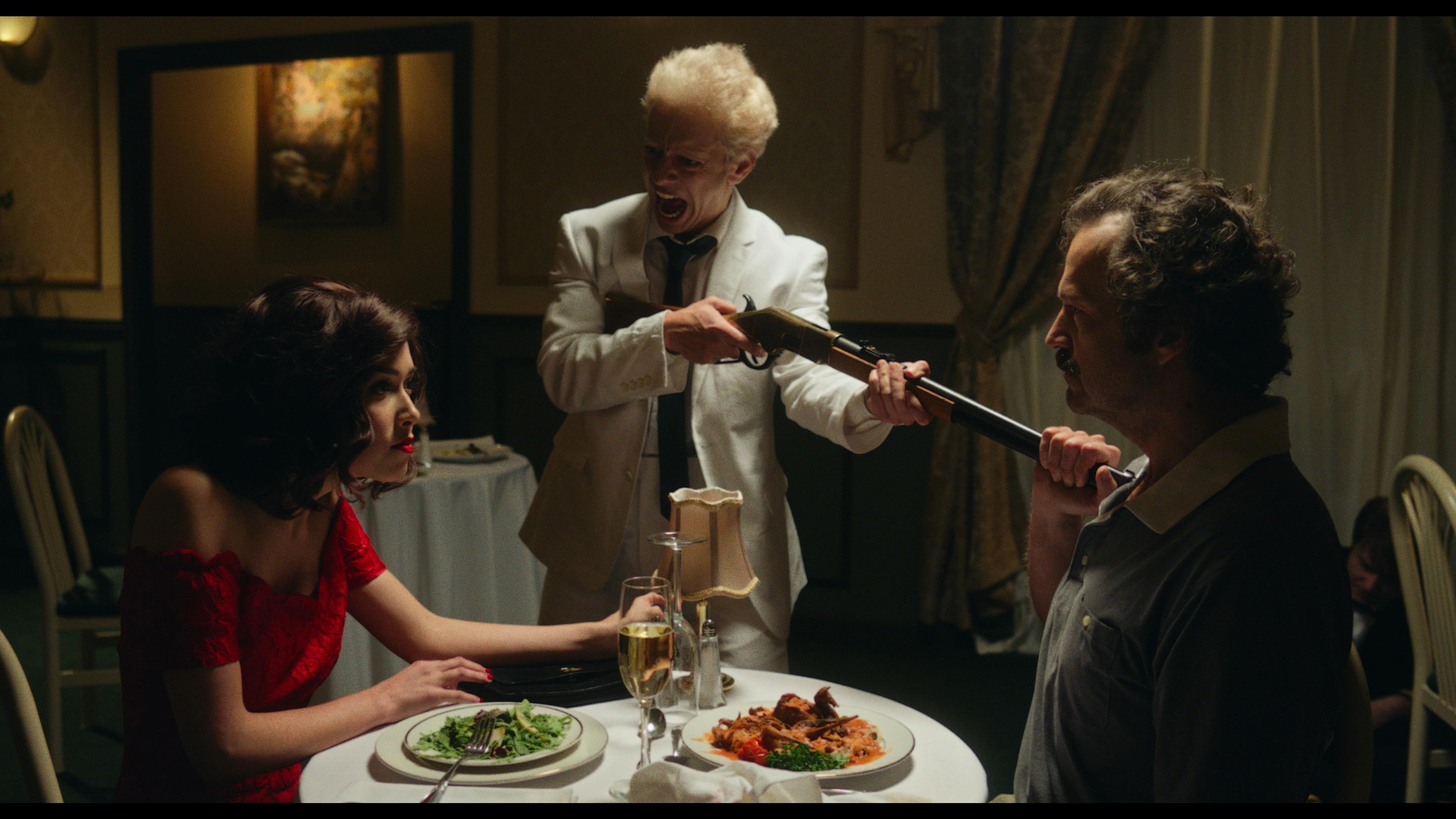 A man in white points a shotgun at a man sitting at dinner with a woman in a red dress