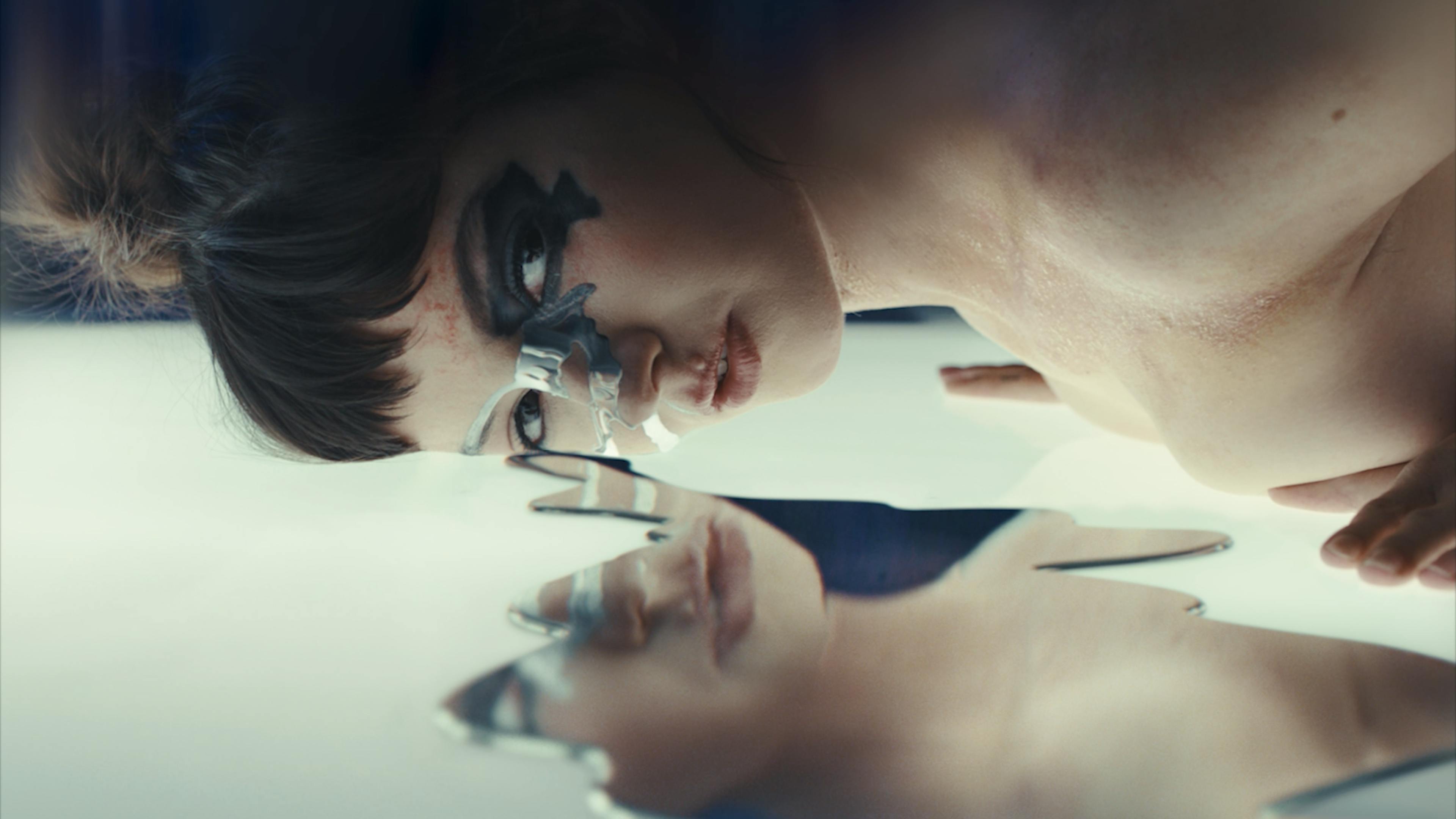 A woman lays on a clear floor with metallic liquid reflecting her image