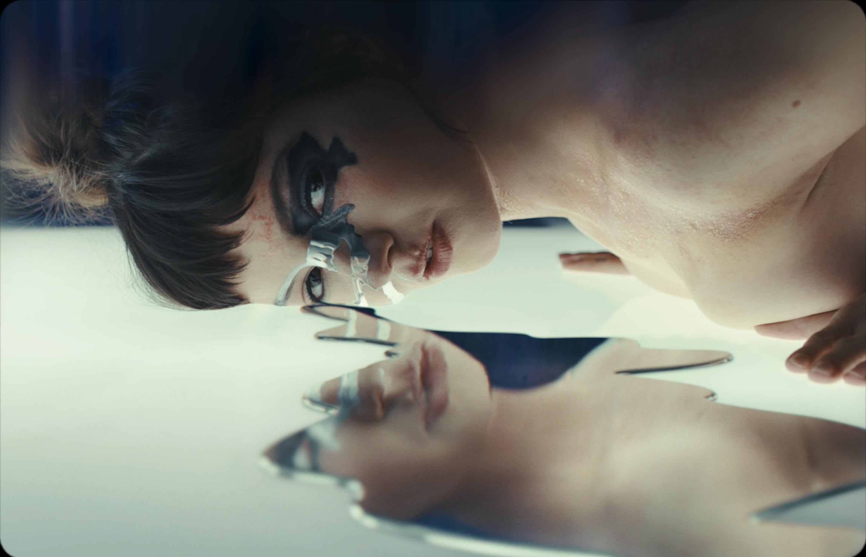 A woman lays on a clear floor with metallic liquid reflecting her image