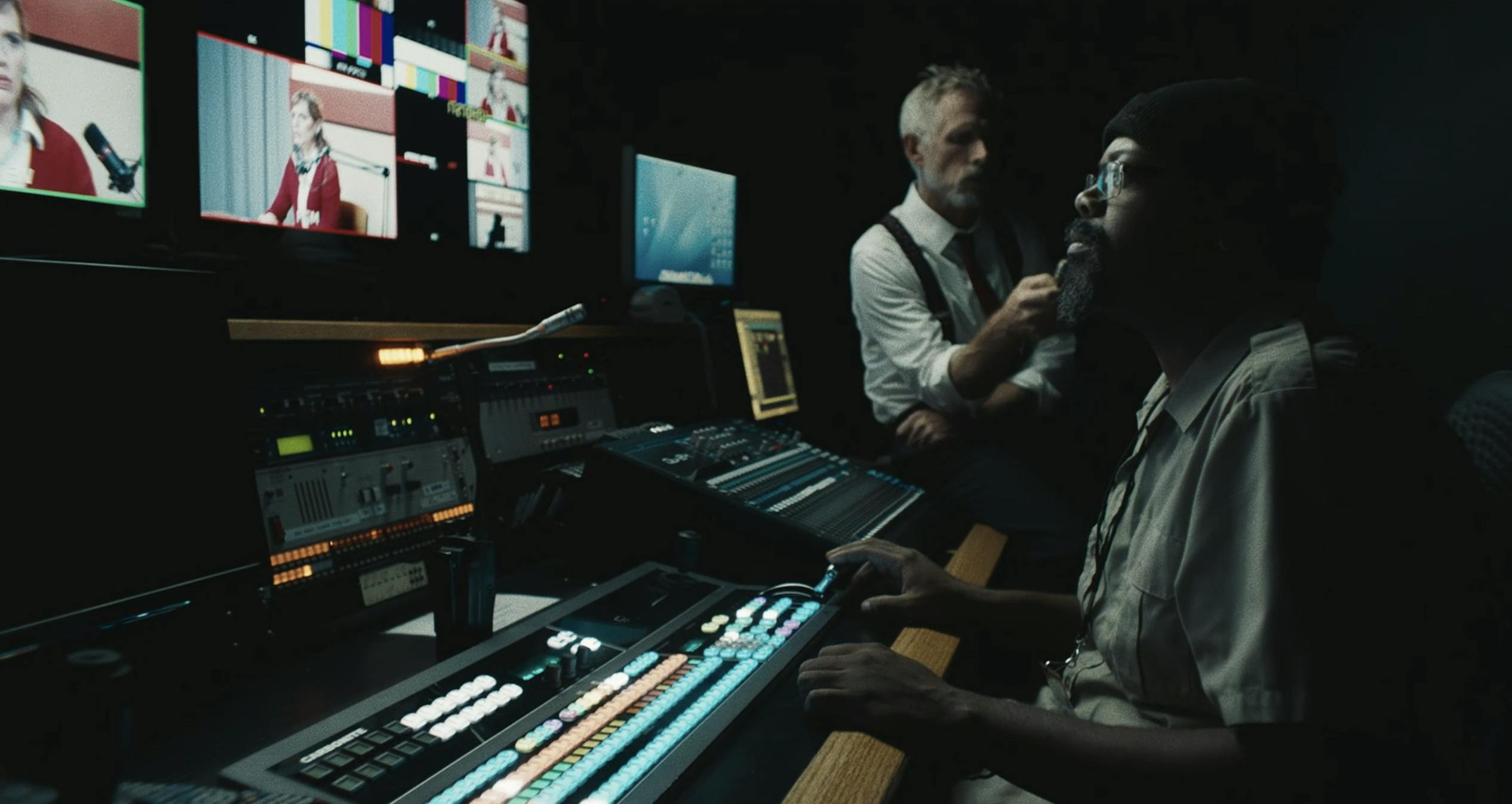 Two men sit in a broadcast studio watching a newscaster on multiple screens