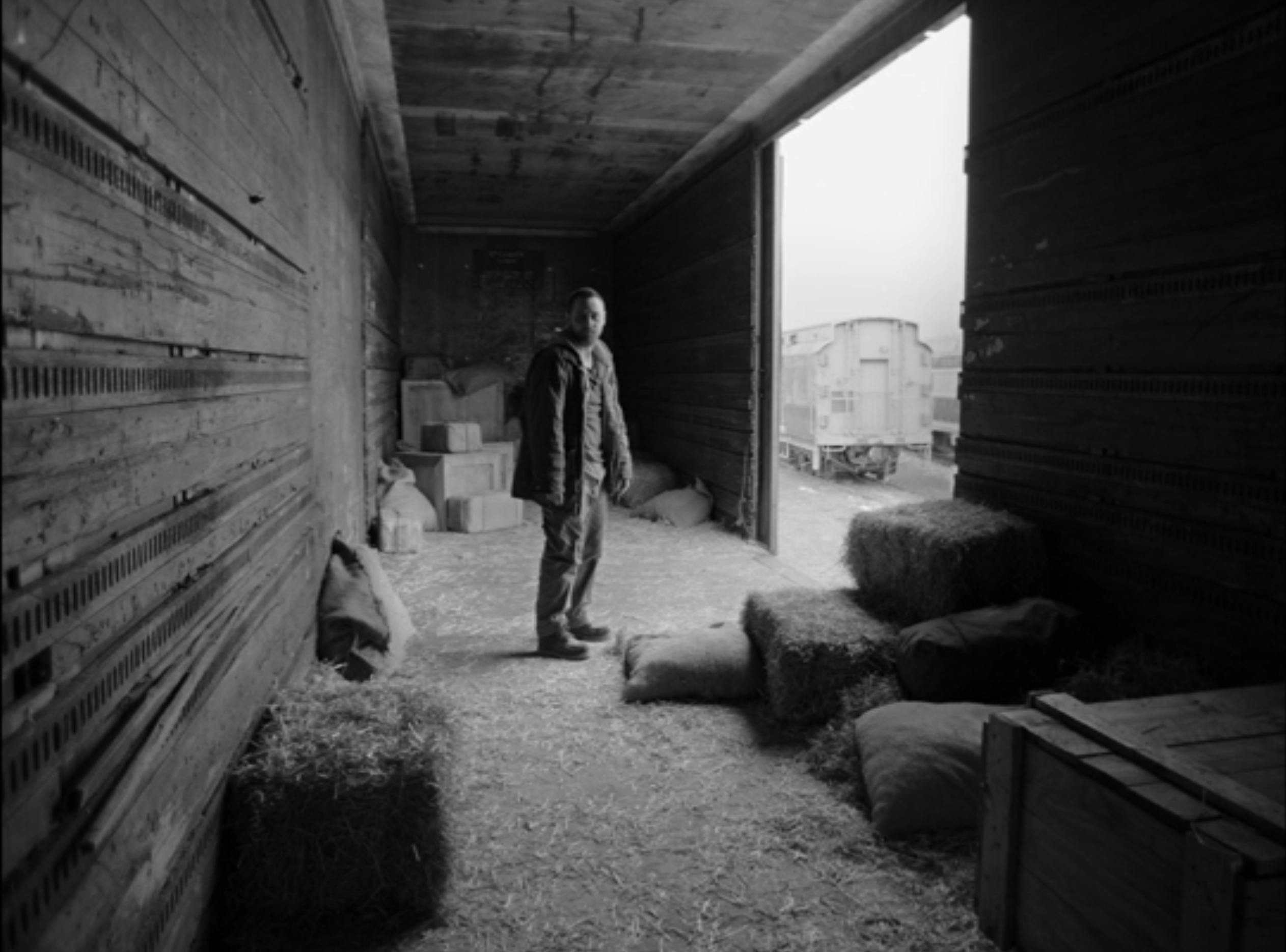 A black and white image of a man in a room with hay stacks and boxes while a railcar is seen outside