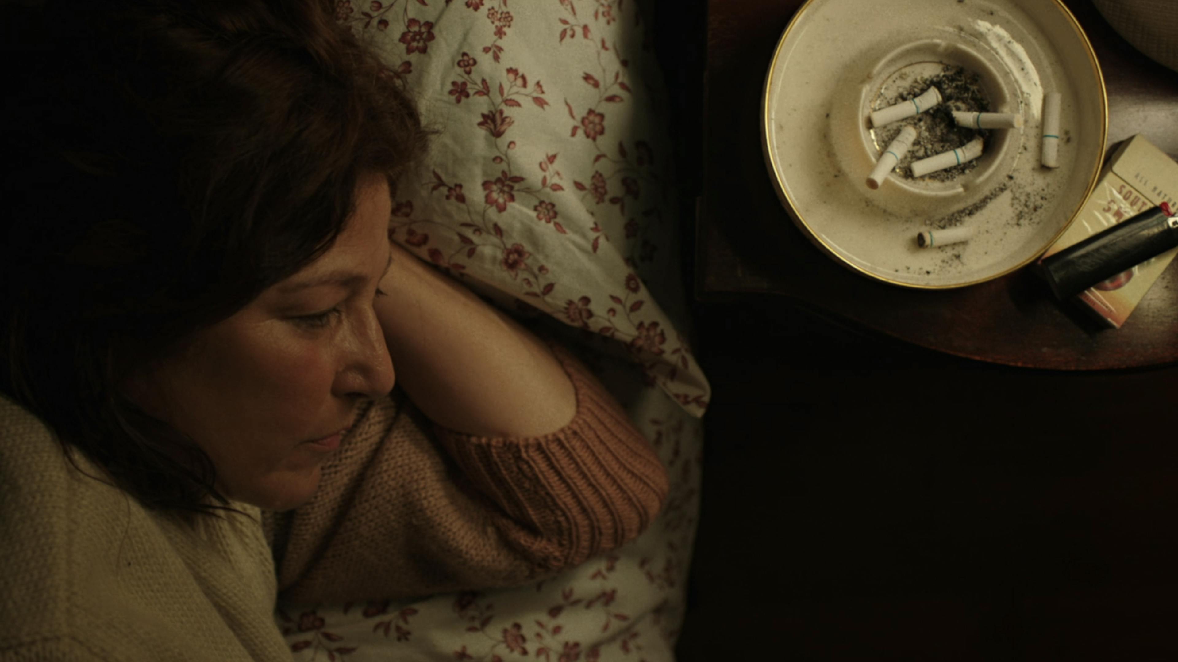 A woman lies in bed next to an ashtray with cigarettes