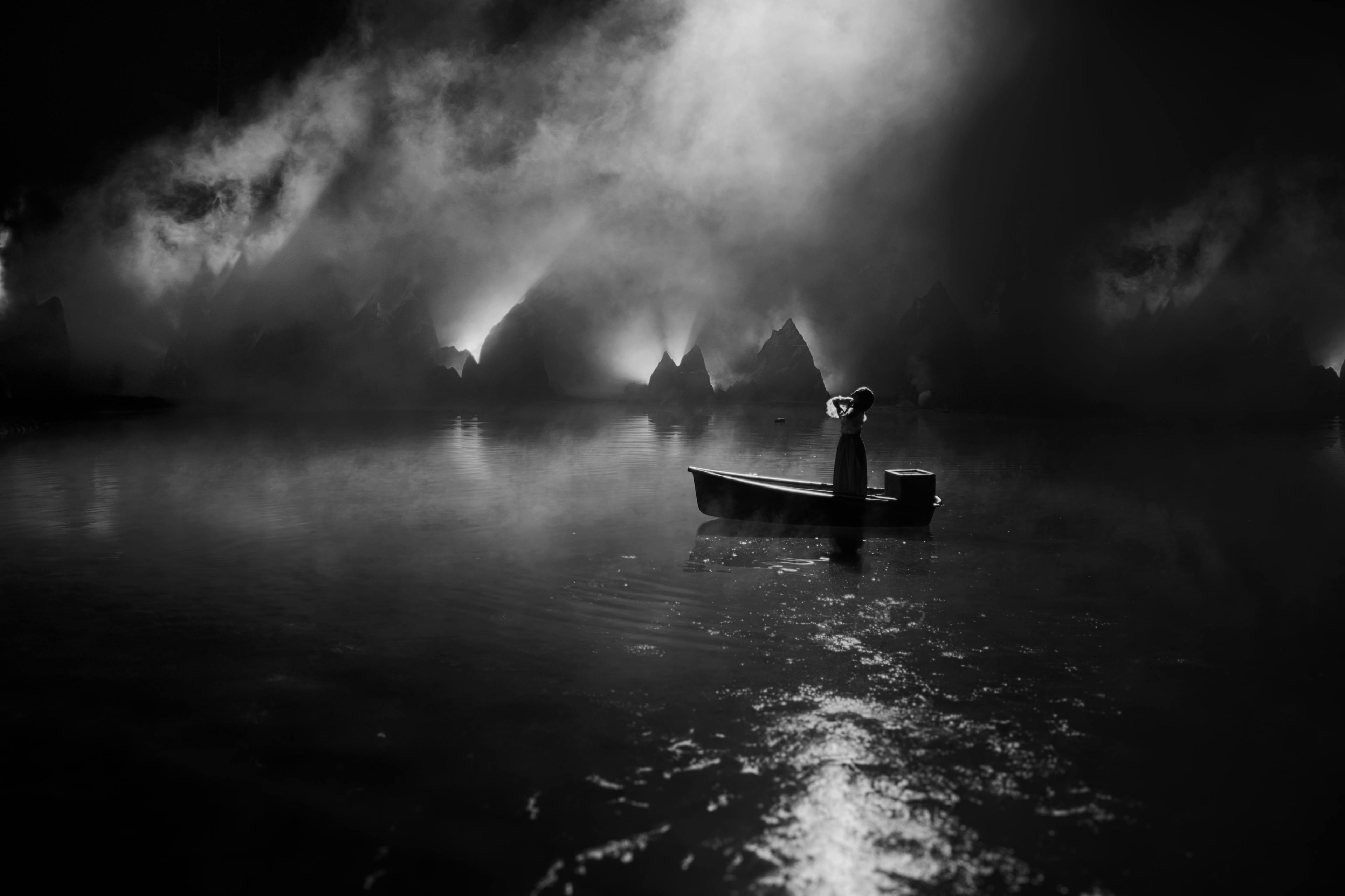 Mitski in a wooden row boat in the middle of a body of water with fog and mountains in the back
