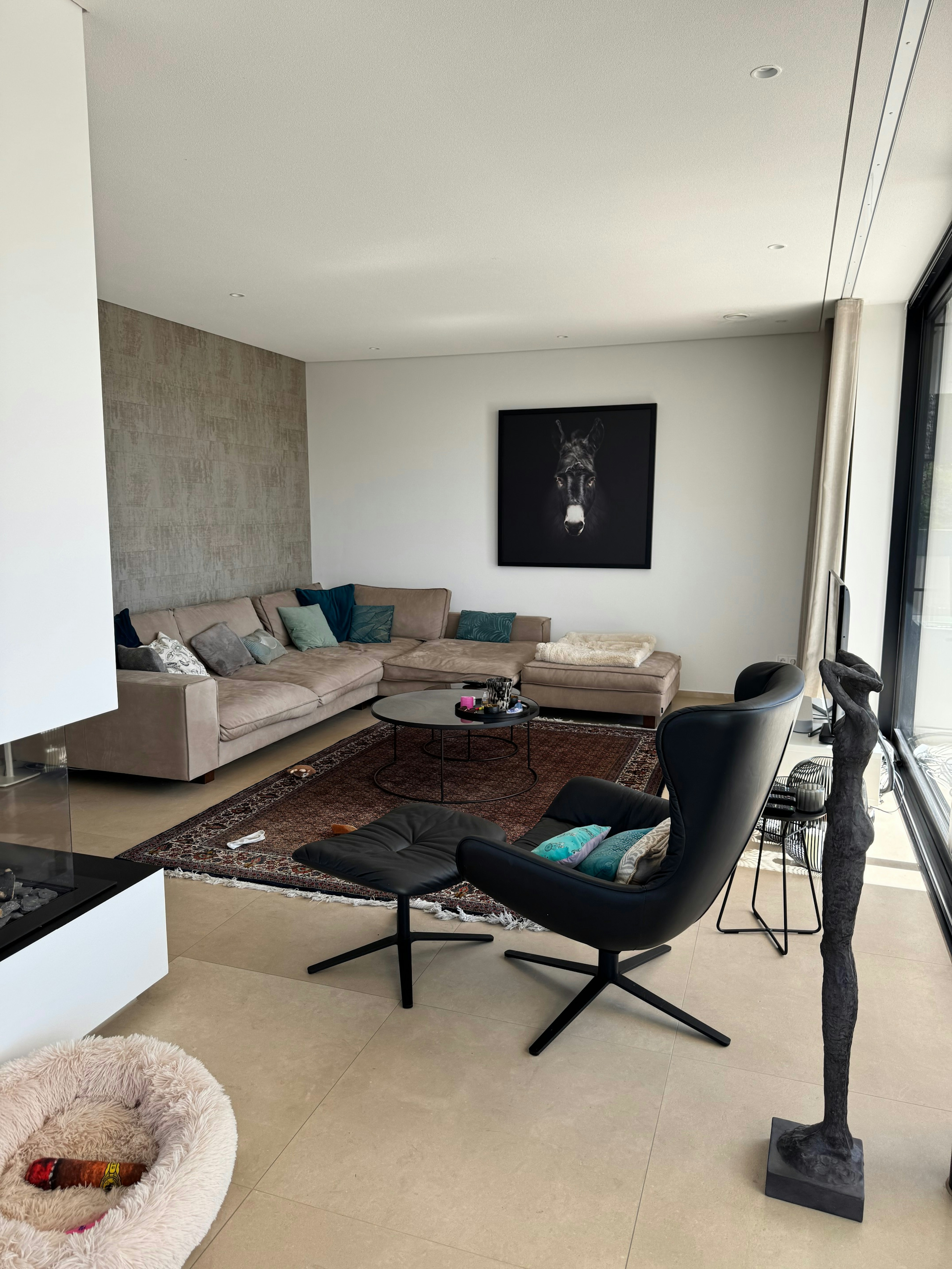 Gemütliches Wohnzimmer mit heller Eckcouch, Kunst an der Wand und großem Teppich – moderne Wohnraumgestaltung mit Blick ins Grüne.