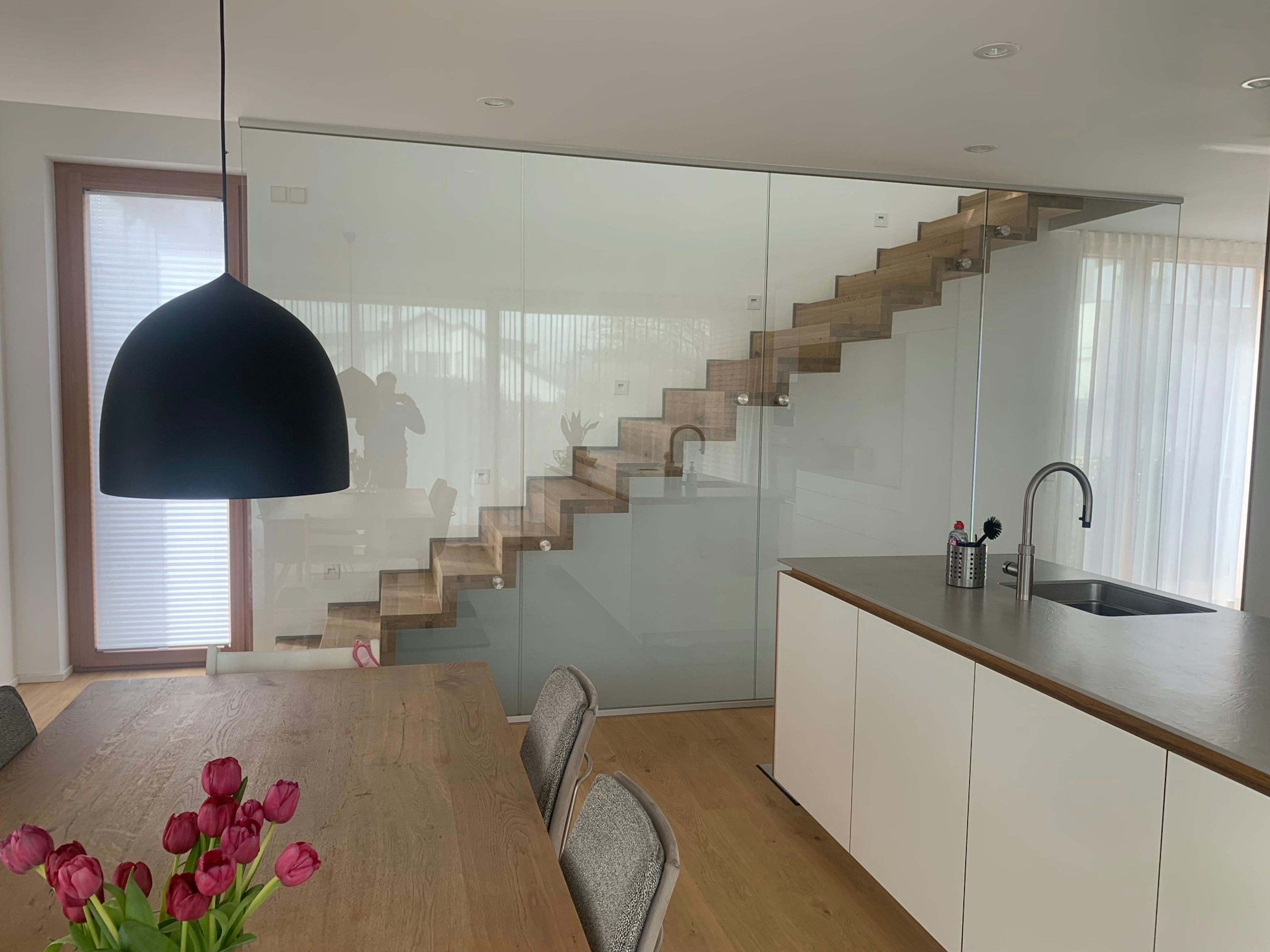 Innenansicht mit Essbereich und freitragender Holztreppe hinter Glas – klare Raumstruktur mit moderner Küchennische.