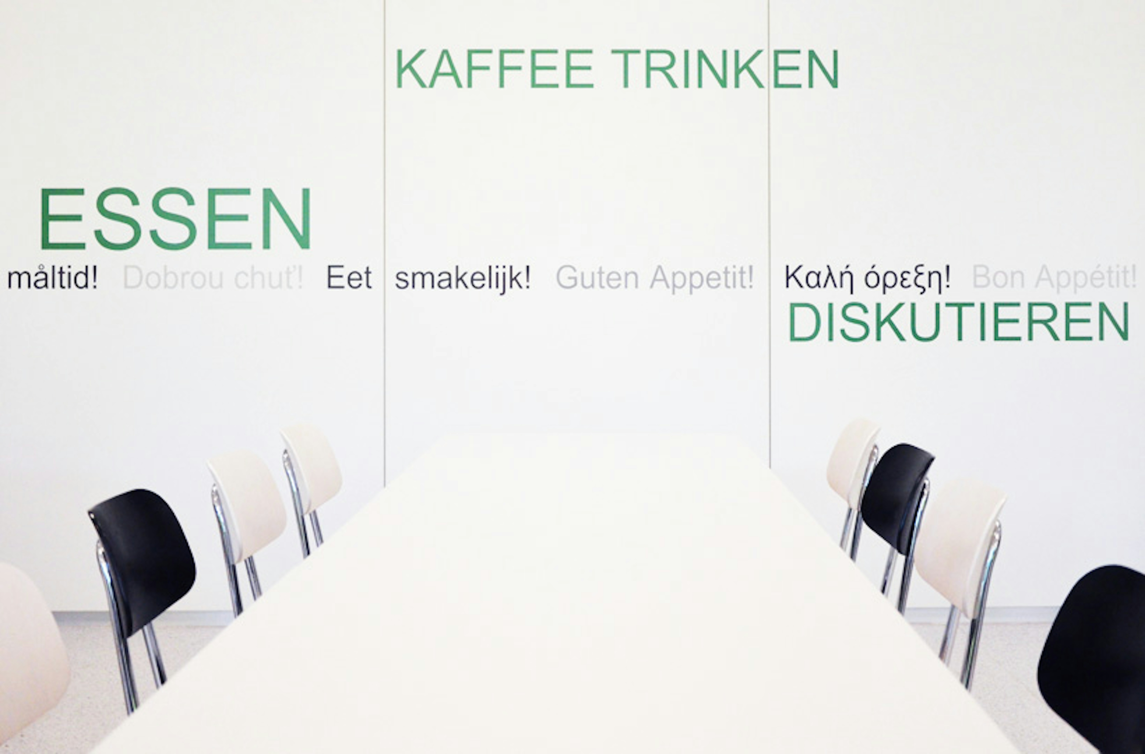 Detailansicht der Mensa mit klarer, reduzierter Gestaltung – weiße Tischgruppe vor typografischer Wand mit mehrsprachigem Guten-Appetit-Schriftzug.