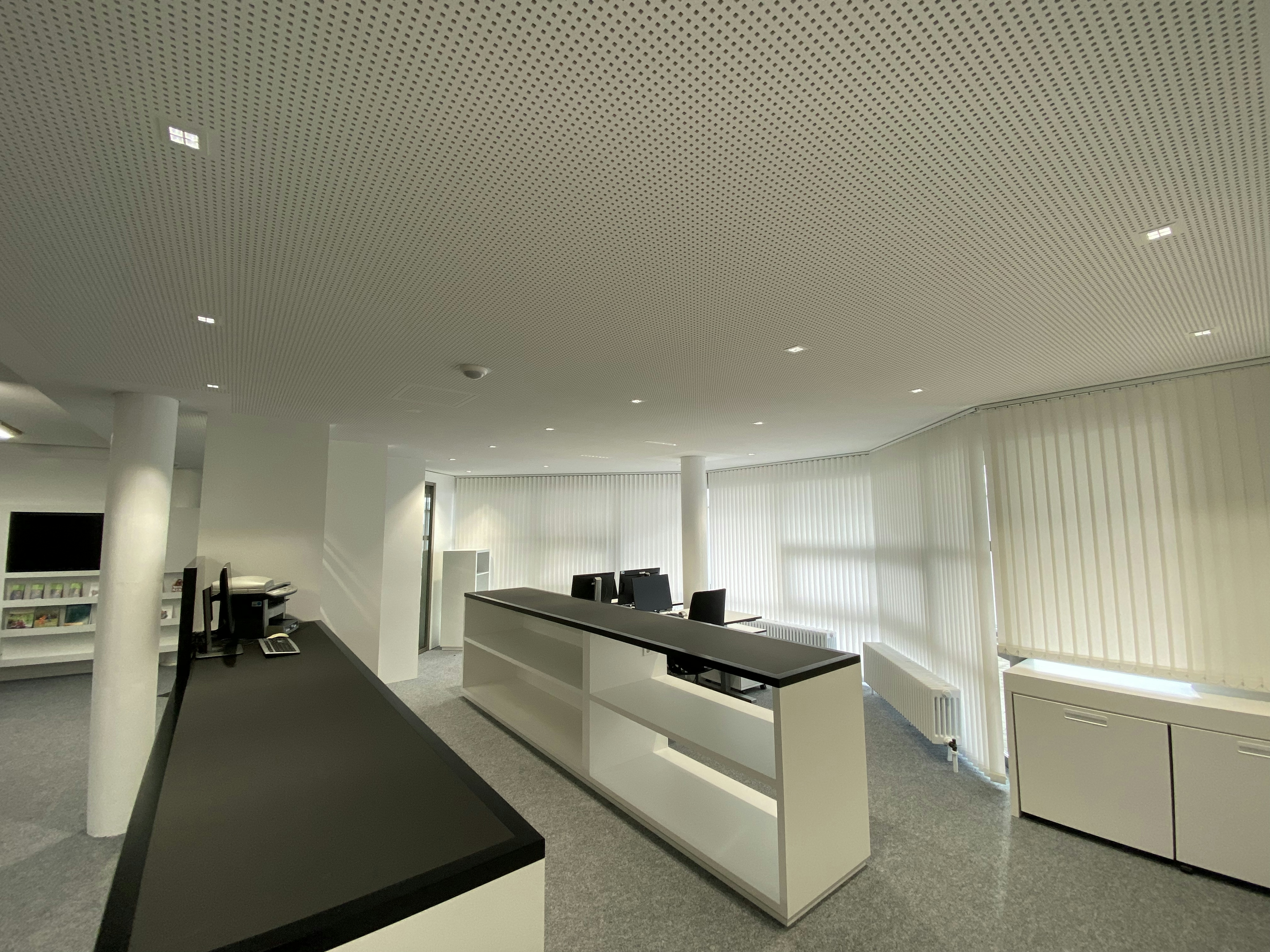 Lichtdurchfluteter Büroraum mit weißen Vertikaljalousien, offenen Regalen und minimalistischer Einrichtung.