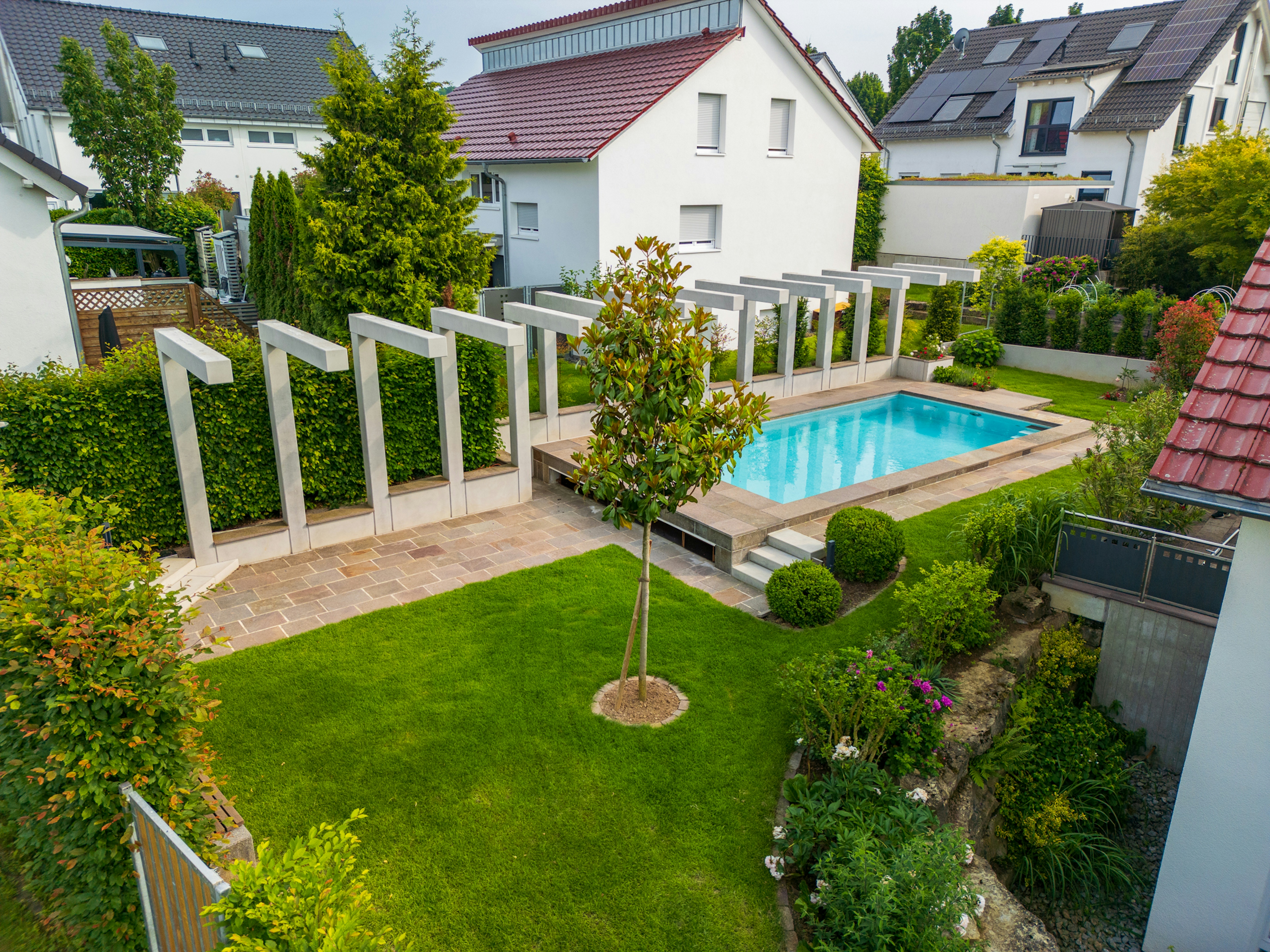 Gartenansicht mit zentralem Baum, Sichtbeton-Pergola und Pool, eingebettet in eine dichte Wohnumgebung