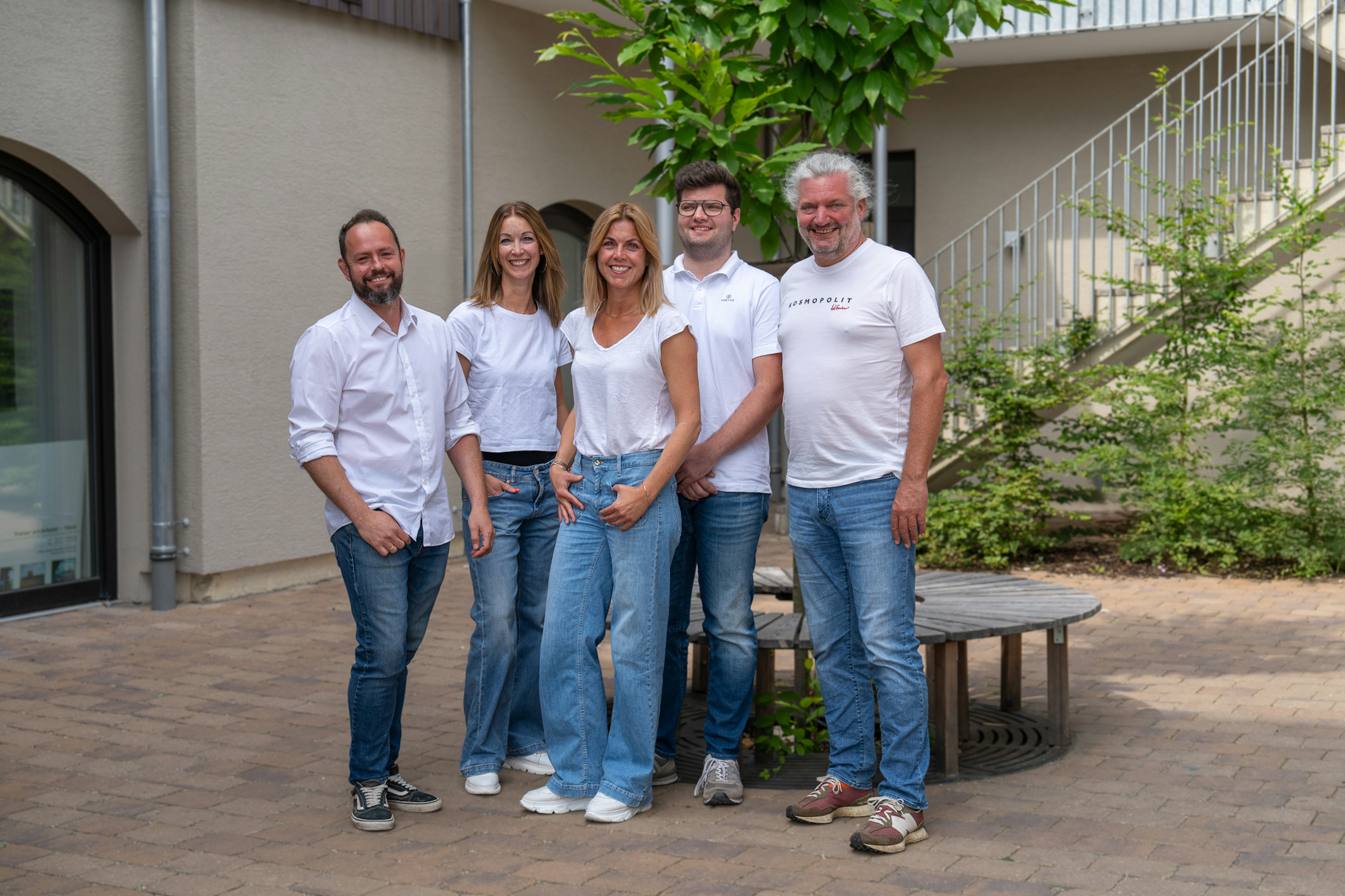 Team des Architekturbüros Bauer stehen lächelnd im Innenhof, alle in weißem Oberteil und Jeans gekleidet – das Team wirkt offen, freundlich und nahbar.
