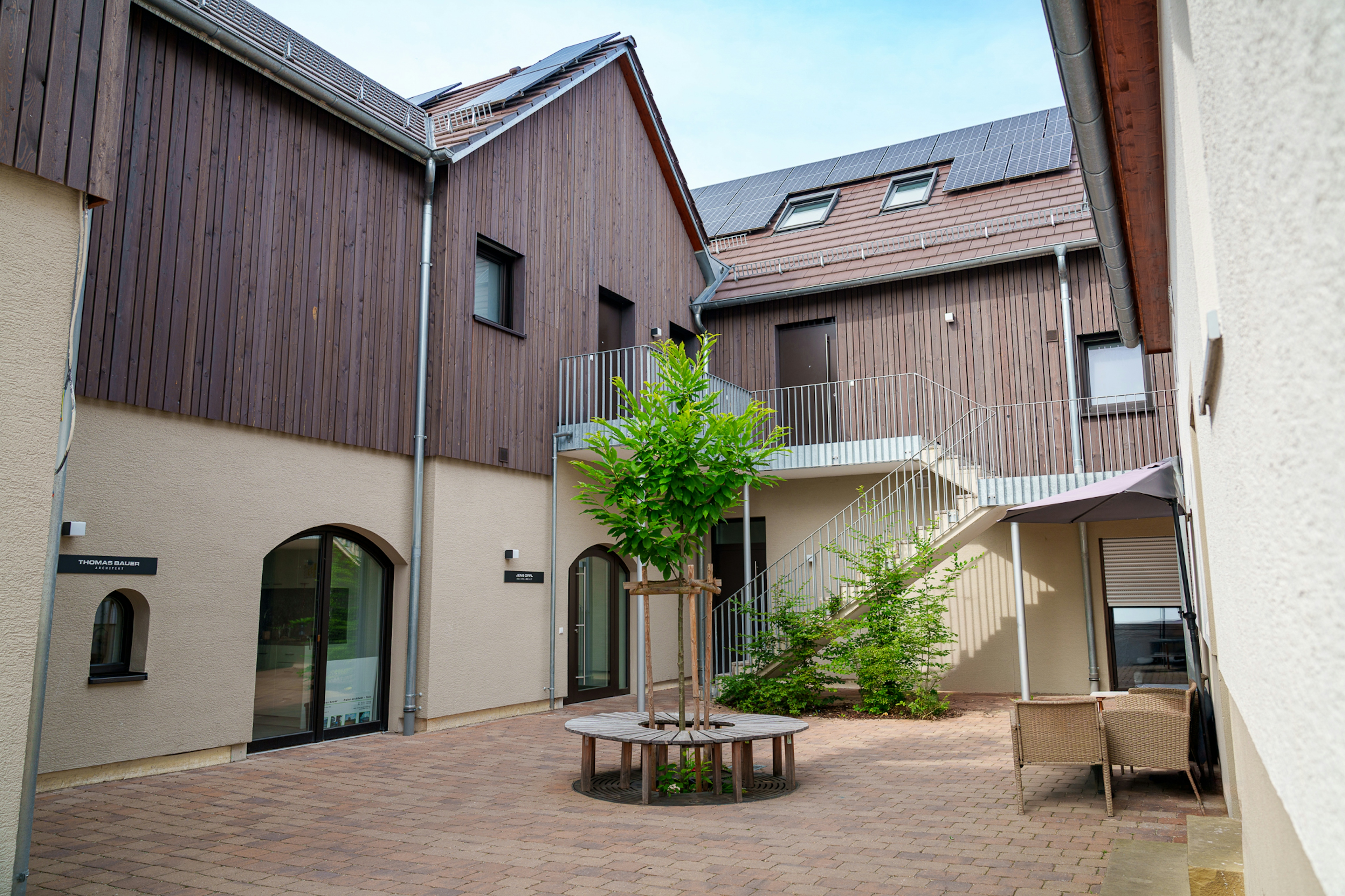 Innenhof des sanierten Weinguts mit junger Bauminsel, Rundbank, Natursteinpflaster und Zugang zu den Büroeinheiten.