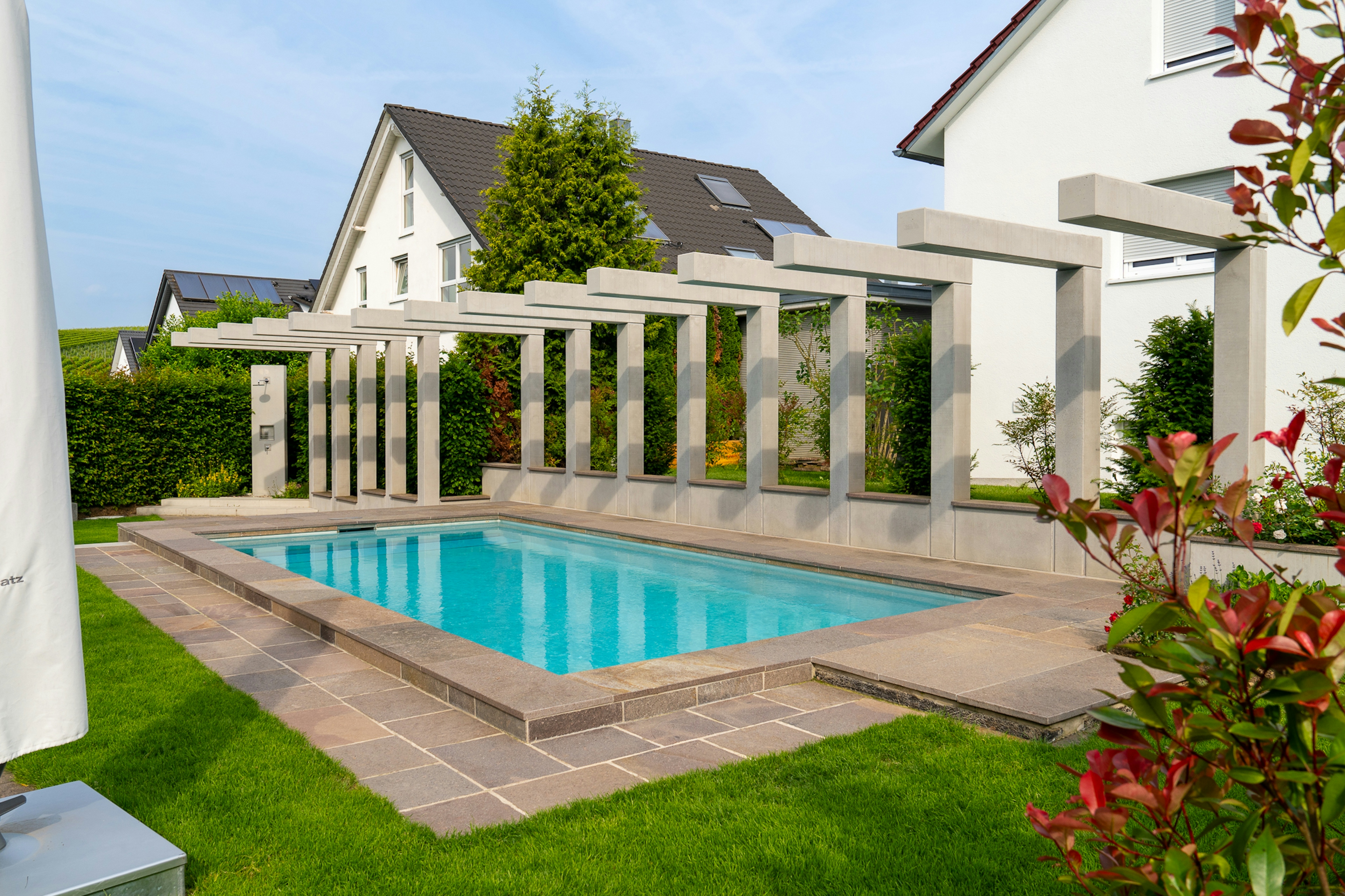 Schwimmbecken mit umlaufender Steinverkleidung, flankiert von Sichtbeton-Pergola, moderner Wohnhaus-Hintergrund