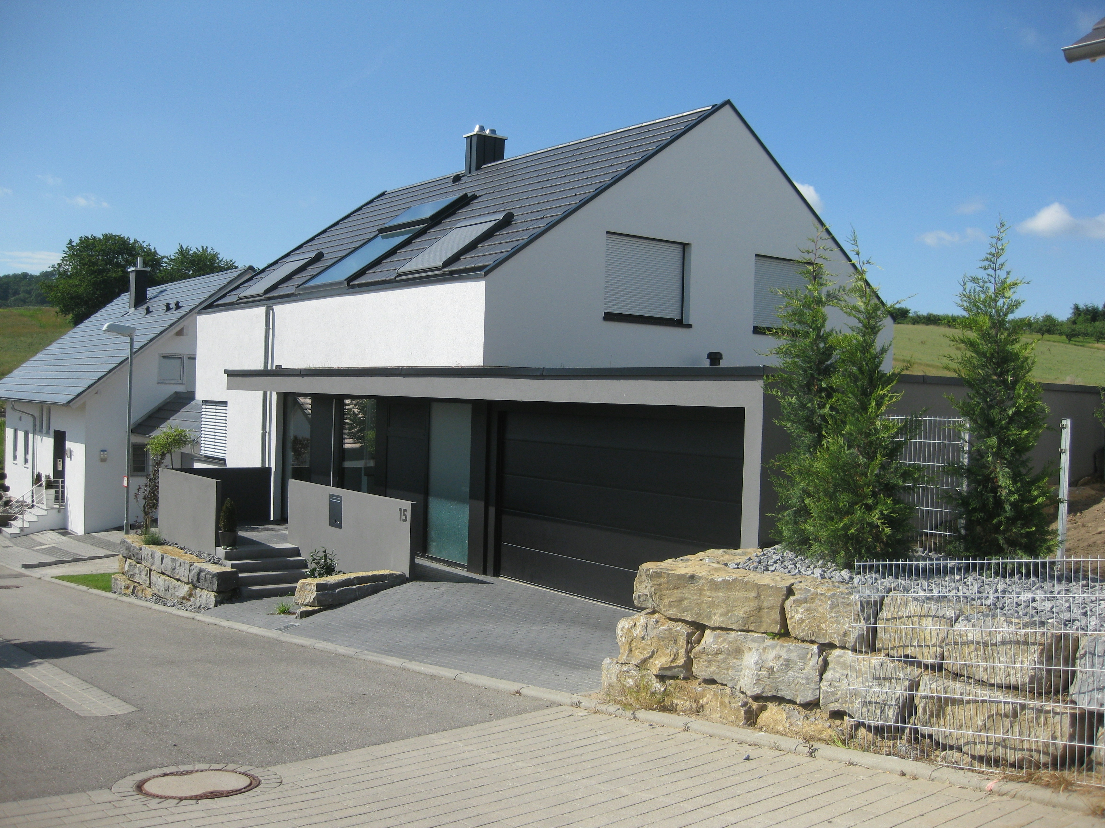 Modernes Einfamilienhaus mit weißer Fassade, dunklen Fensterrahmen und klarer Kubatur, eingebettet in eine grüne Umgebung.