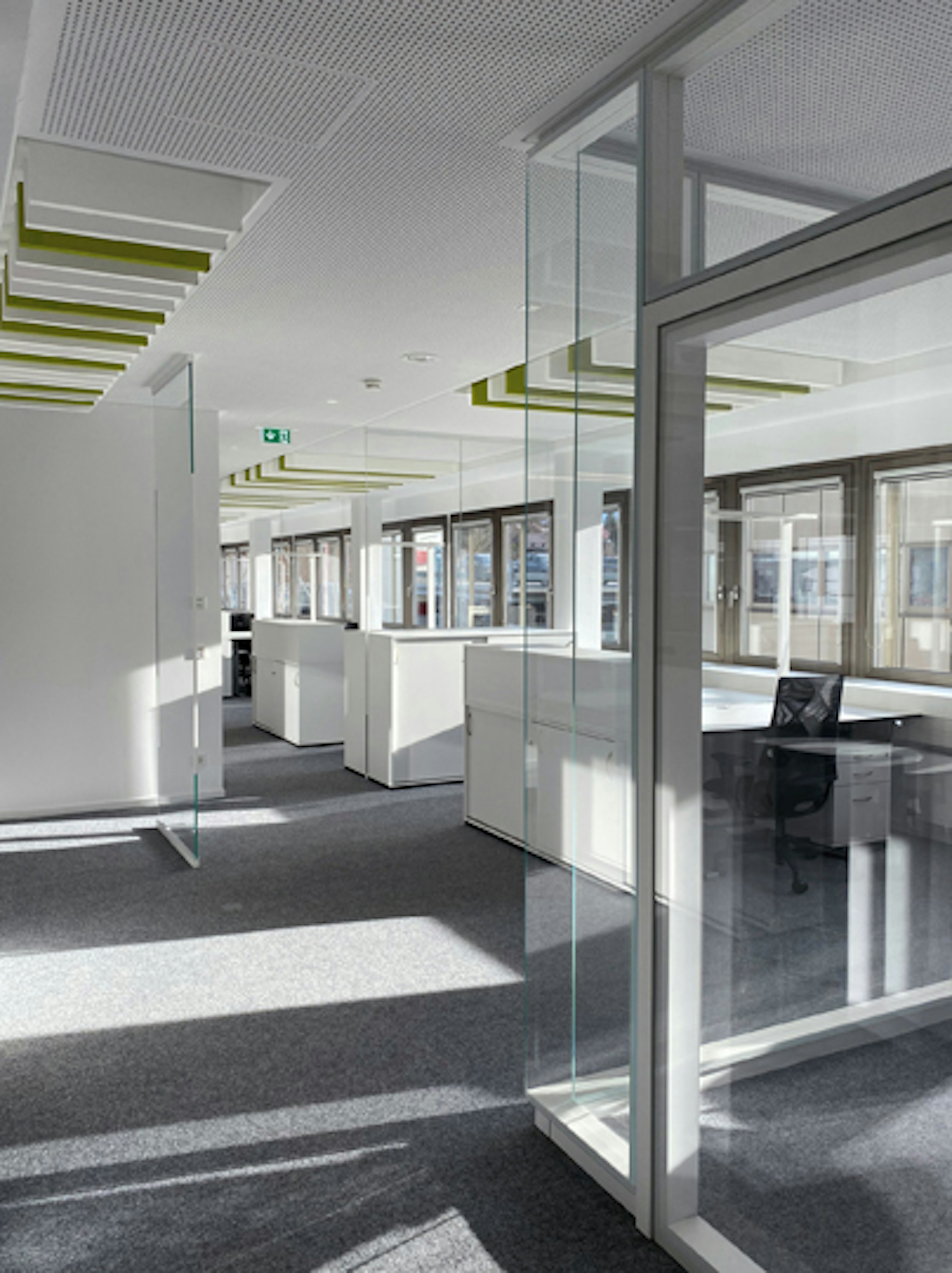 Lichtdurchfluteter Büroflur mit mehreren weißen Arbeitsinseln und grün-akzentuierten Deckenpaneelen.