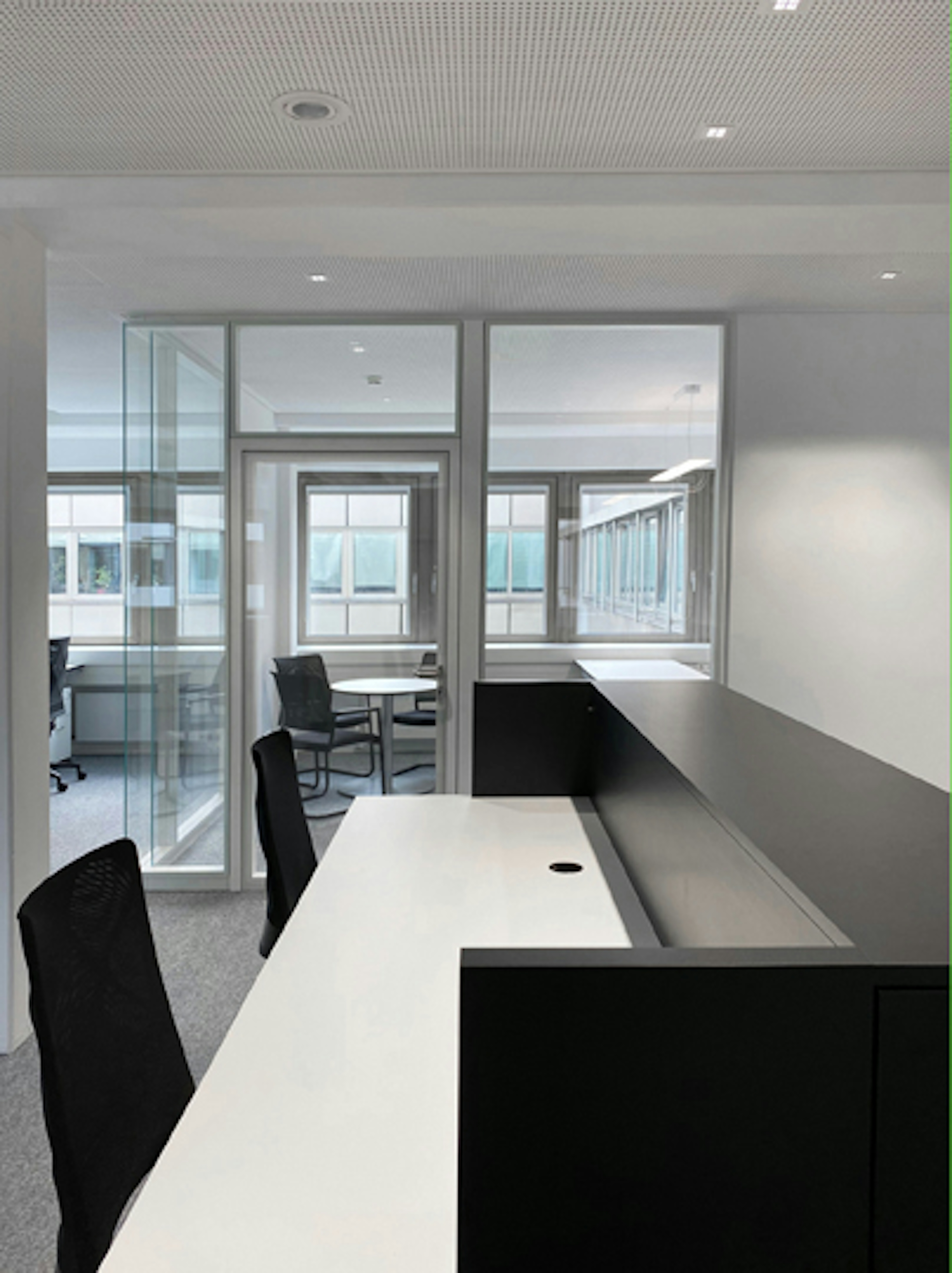 Einzelner Arbeitsplatz mit Sicht auf ein separates Besprechungszimmer mit Rundtisch und zwei Stühlen, umgeben von Glaswänden.