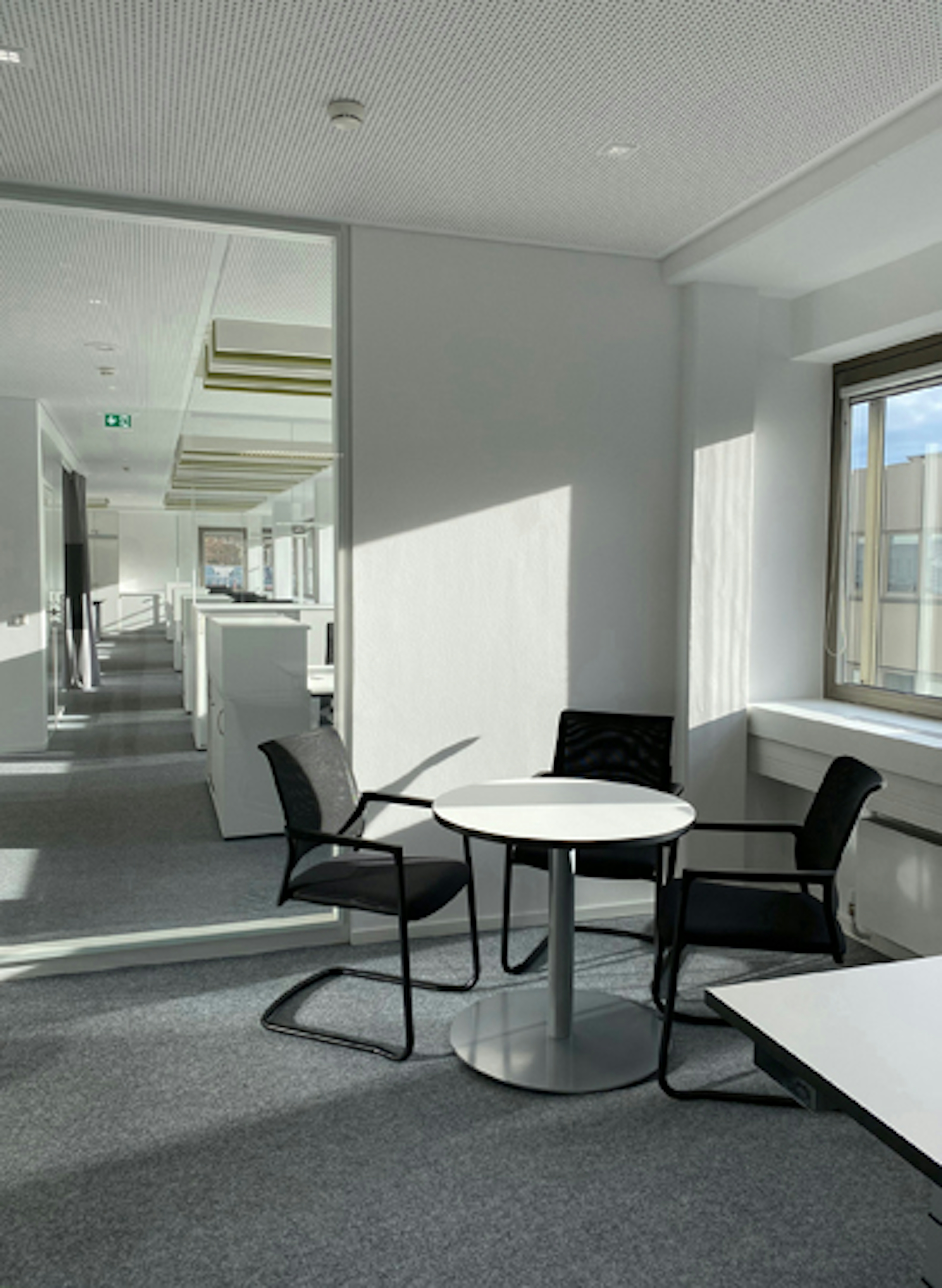 Kleiner Sitzbereich mit rundem Tisch und zwei Stühlen am Fenster, daneben strukturierter Büroflur mit Schränken und Arbeitsplätzen.