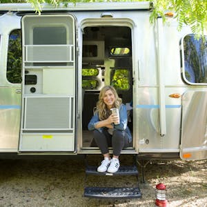 Image of Sarah Schulz sitting in the doorway of an Airstream travel trailer RV, smiling at the camera while holder a coffee cup.