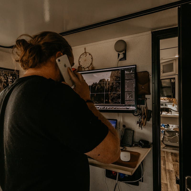 A man standing at a desk, taking a phone call while working on his computer.