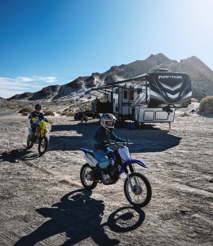 Two people ride dirt bikes in front of a toy hauler rv.