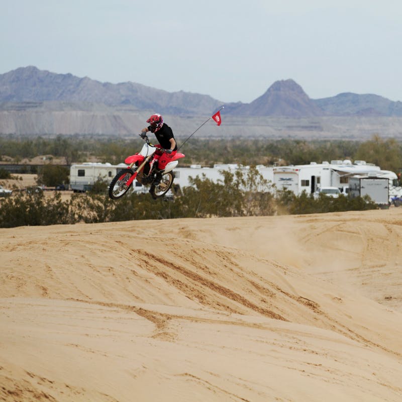 A dirt biker getting air over a sandy dune with RVs in the distance.