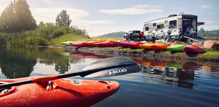 Toy hauler RV on lake edge with a line of kayaks, unloading many colorful kayaks