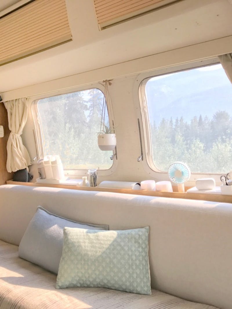 Interior shot of an RV, with cream colored couch, hanging plants and beige curtains, and pine trees outside the window.