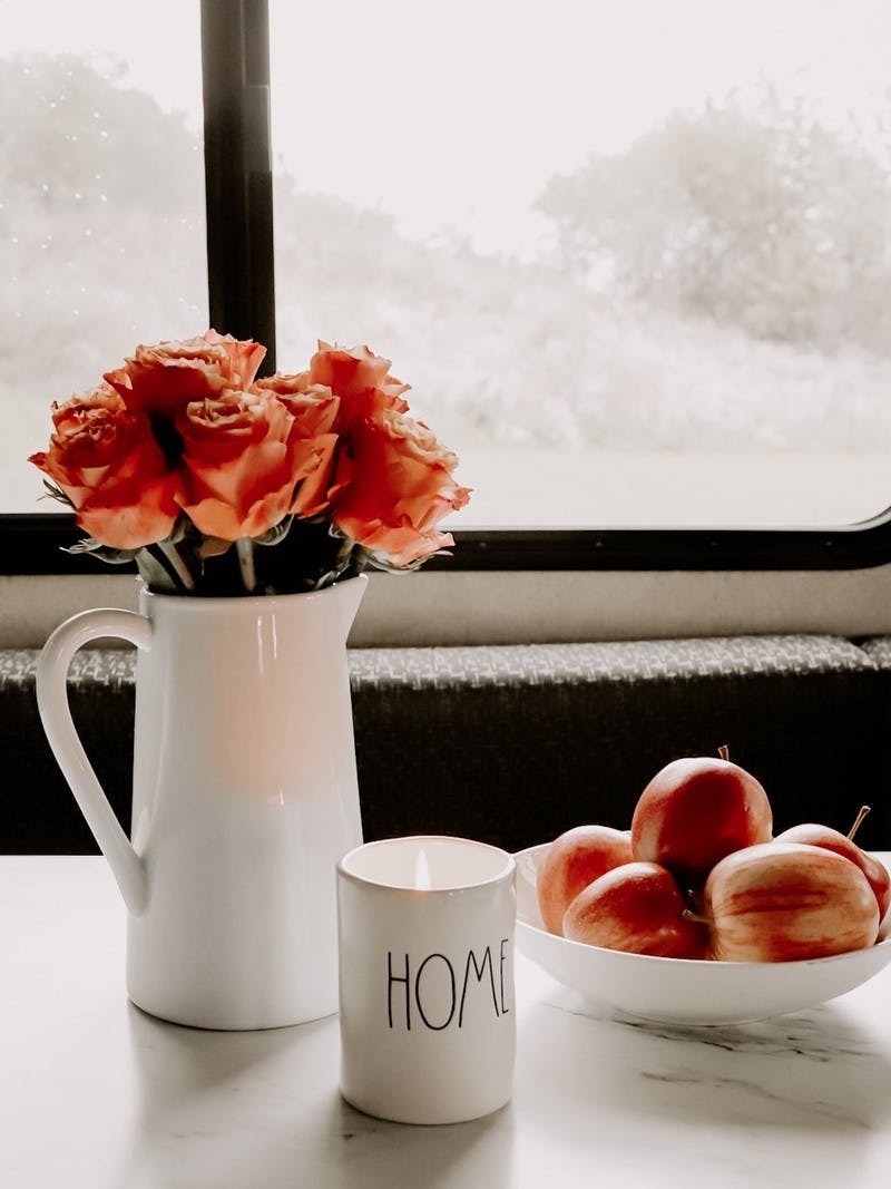 A vase of roses, a bowl of crisp apples and a glowing candle that reads "Home" sit on an RV table in front of a foggy window to the outdoors.