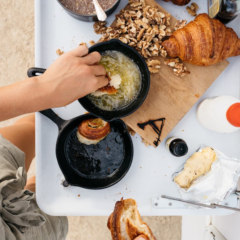 A hand dipping croissant pieces into melted butter and placing them in a small cast iron skillet.