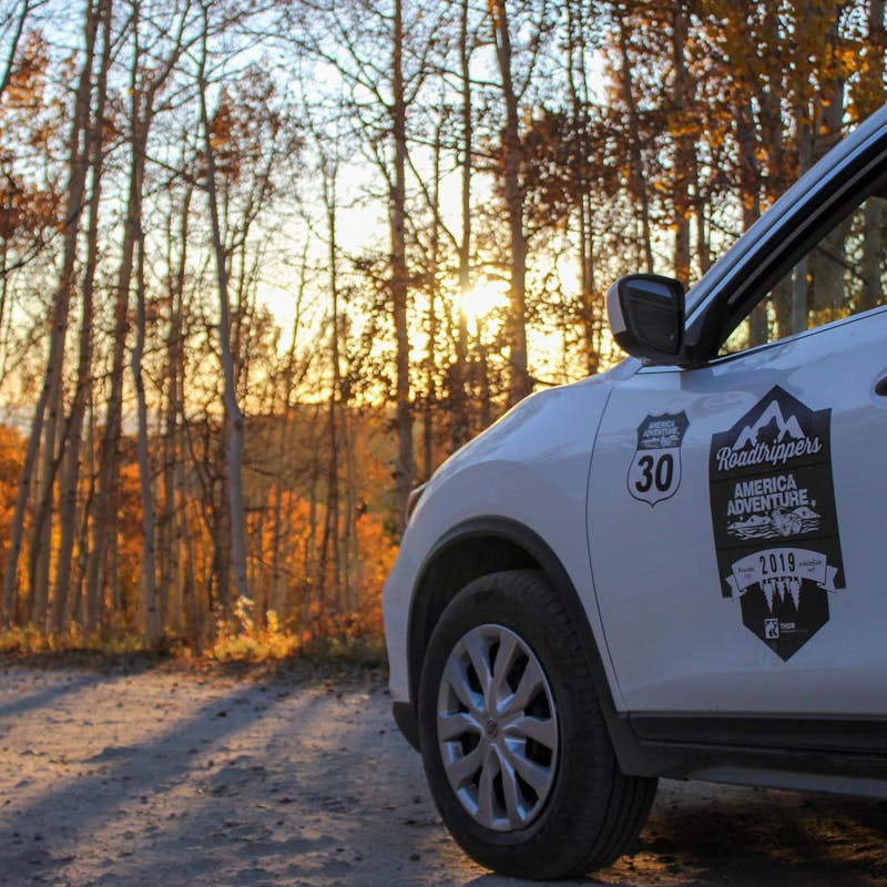 White car with America Adventure decal on dirt road with forest and golden sunset light