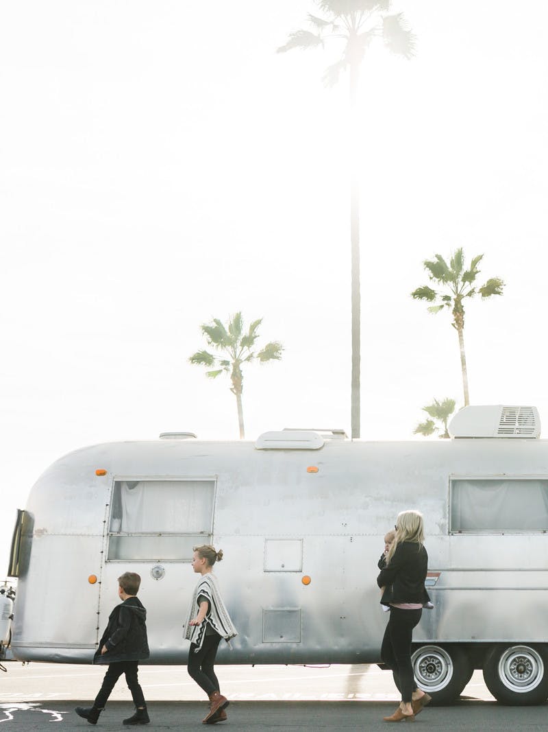 Amber Thrane holding baby, walking in front of Airstream RV, with two kids walking in front and palm trees in the background.