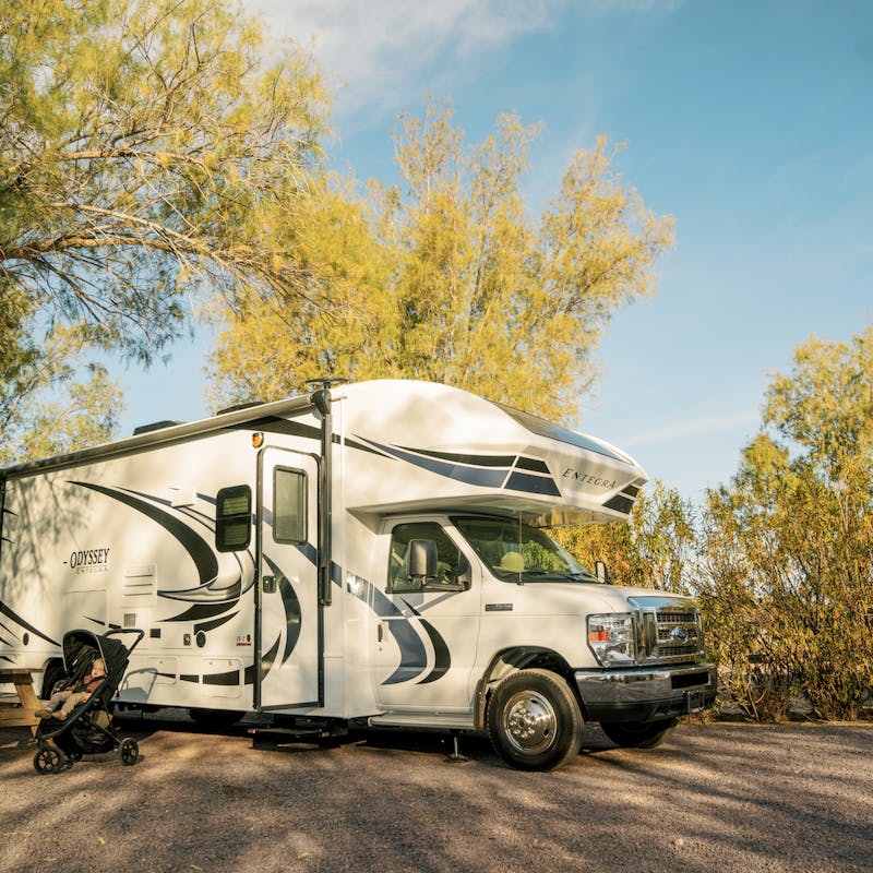 One young boy and child in stroller in front of a Class C motorhome in campground with trees