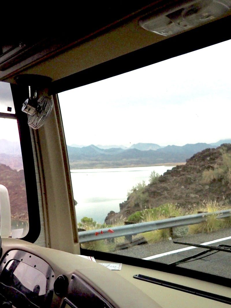 A view of a man driving an RV down the road with a view from inside the RV looking out the front windshield, with a view from the passenger window as well, to see the mountains and ocean.