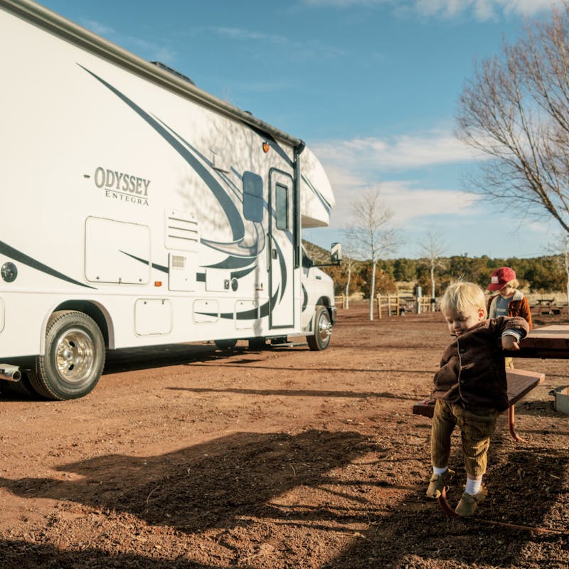 Two young boys sit at picnic table at dirt campsite in front of Thor class c motorhome