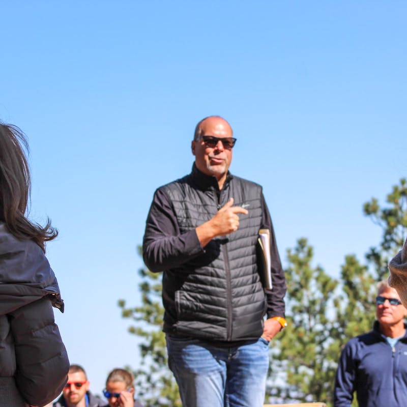Man in black vest and sunglasses speaks to crowd of people outside