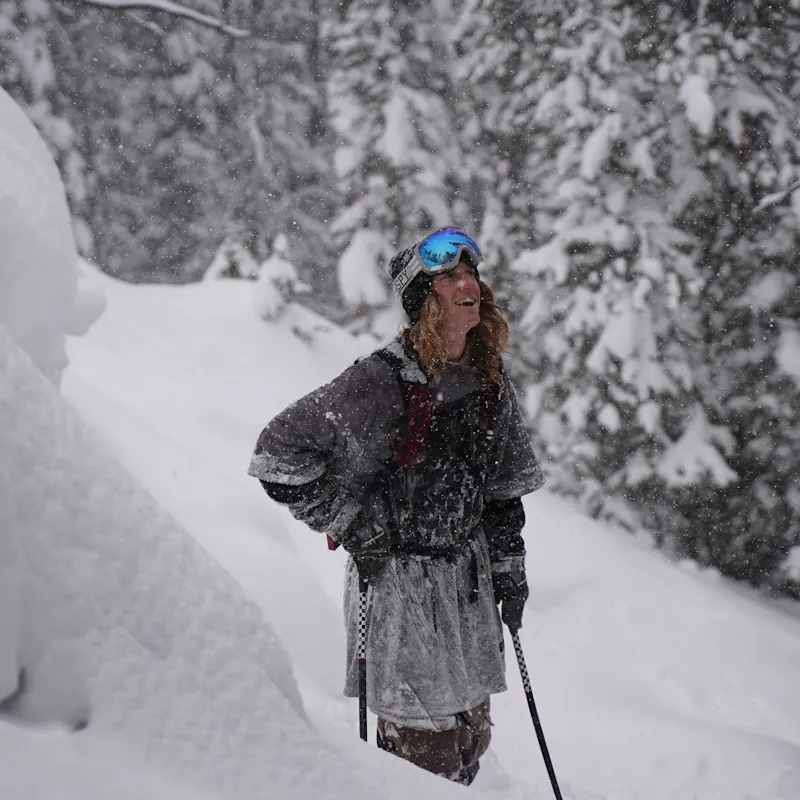 Ryan Barrick with ski poles and ski goggles, surrounded by snow, looking up at the ski and smiling.