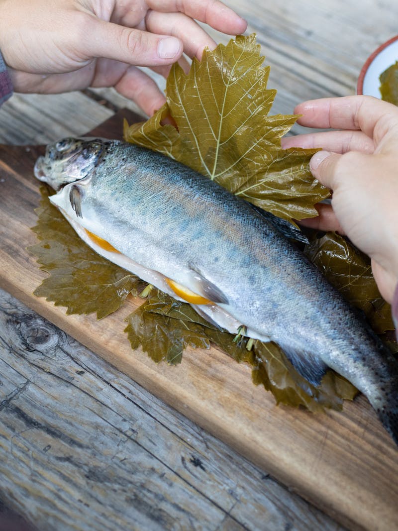 Hands wrapping full, stuffed trout in grape leaves, on a cutting board on a picnic table.
