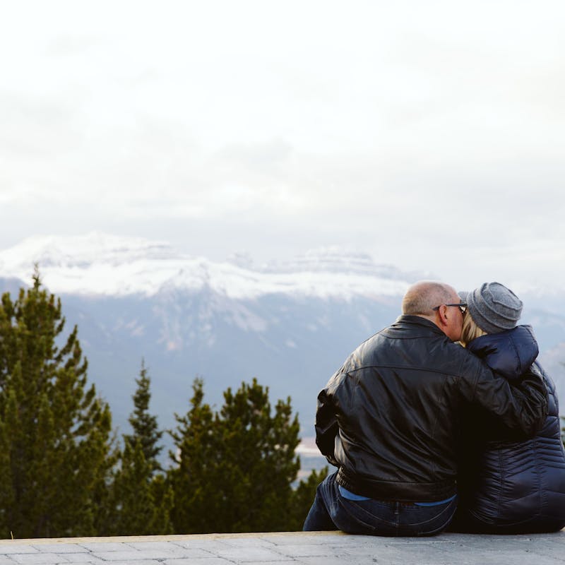 Craig and Tina sitting on a low wall and exchanging a kiss in front of a mountain view.